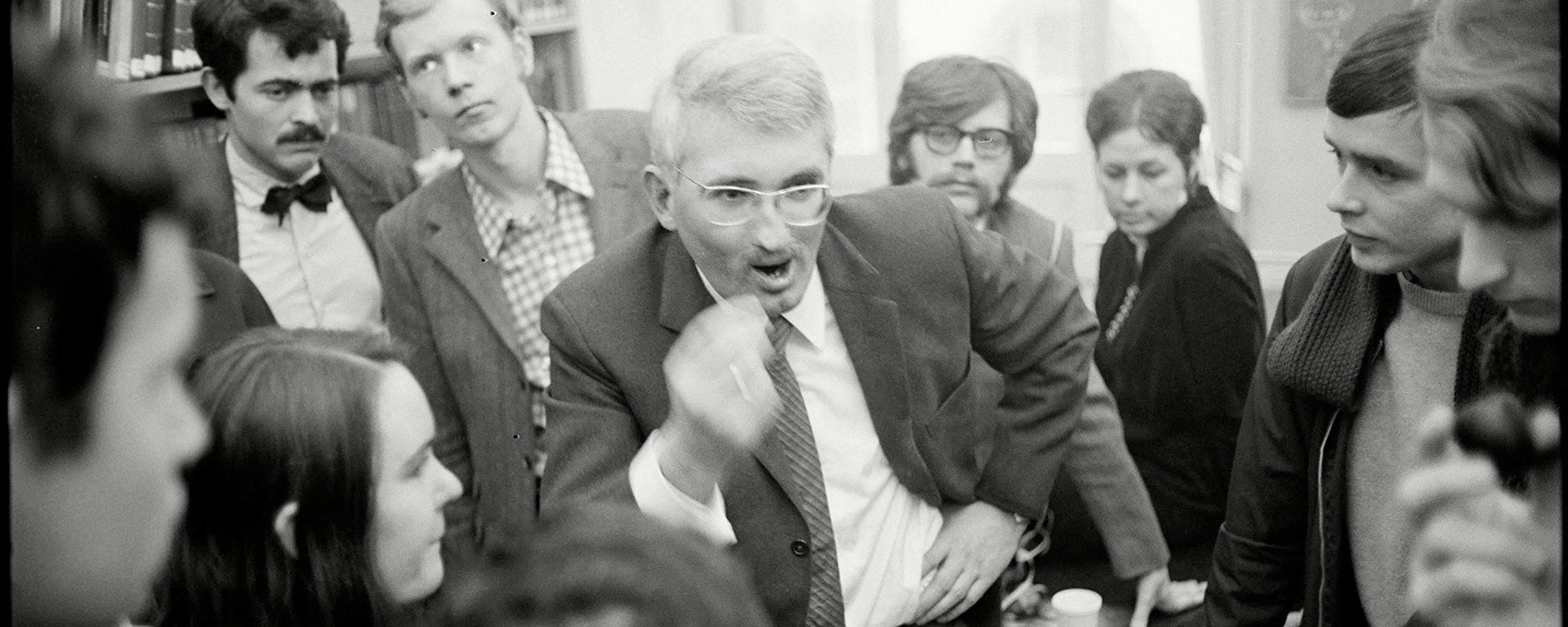 Black and white photo of a man in glasses animatedly speaking to a group gathered around him in an indoor setting.