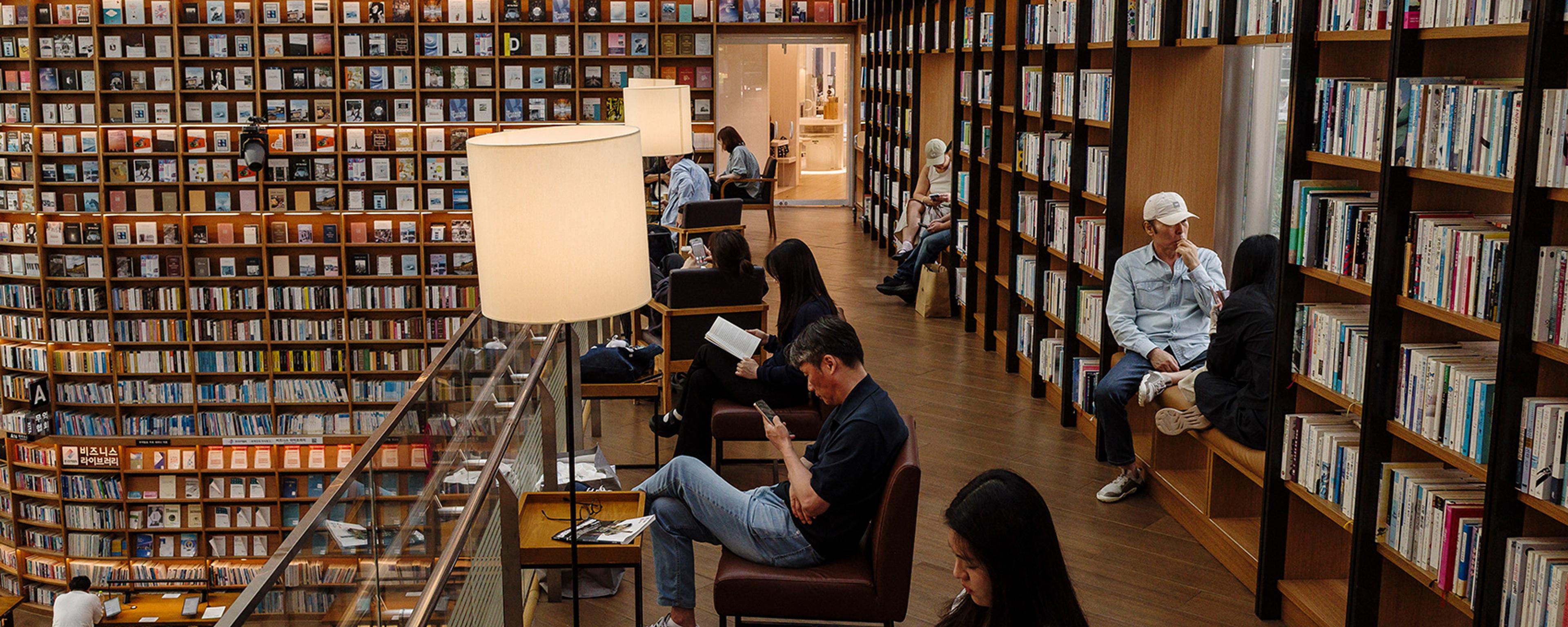 A modern library with tall bookshelves and people reading or using devices in a cosy, well-lit atmosphere.