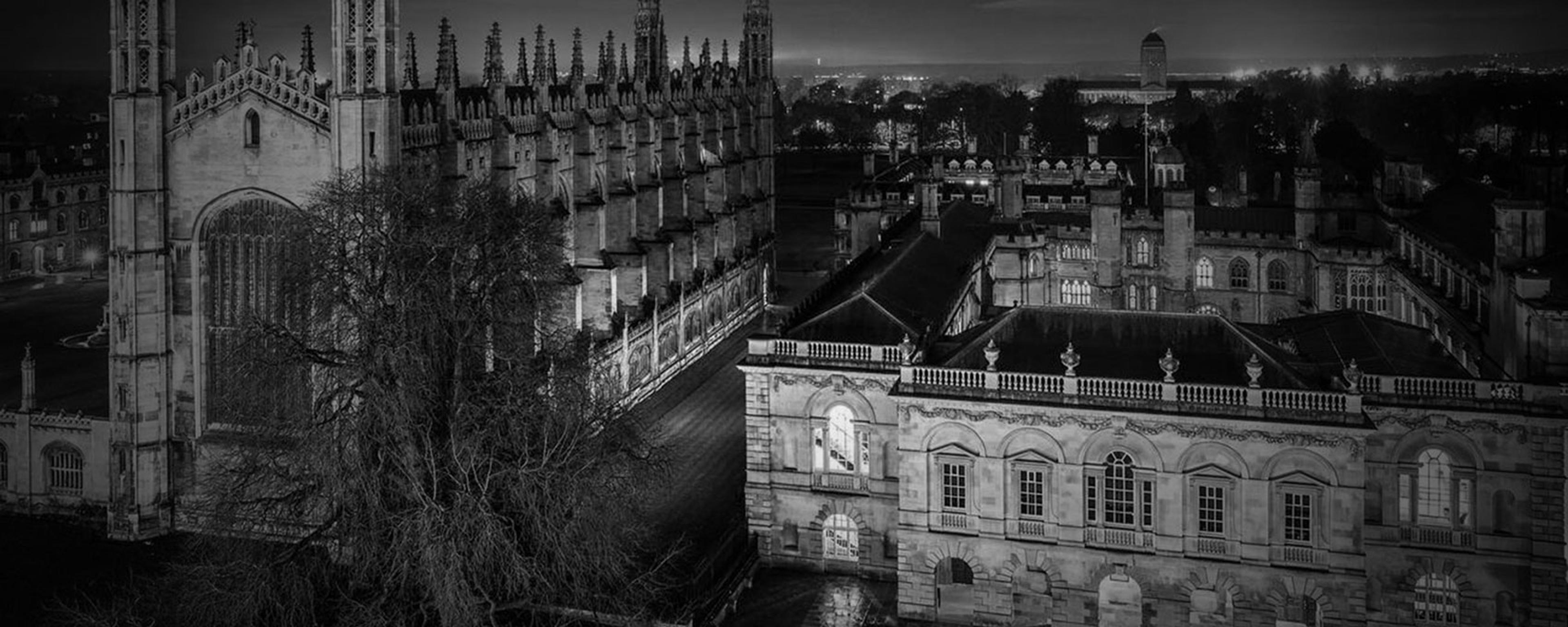 Black and white night photo of historic Gothic-style buildings with arched windows, towers and courtyards.