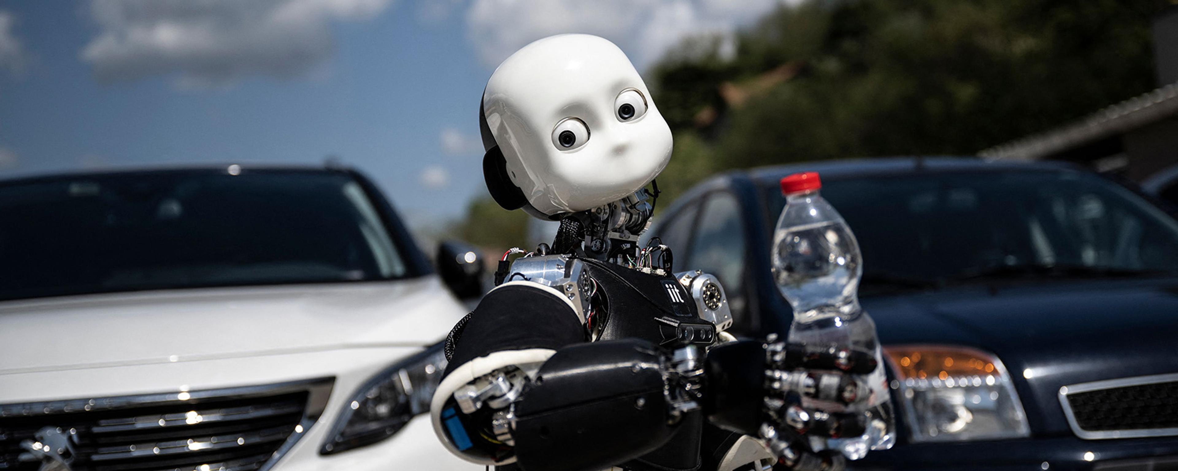 A humanoid robot holding a water bottle in front of parked cars under a partly cloudy sky.