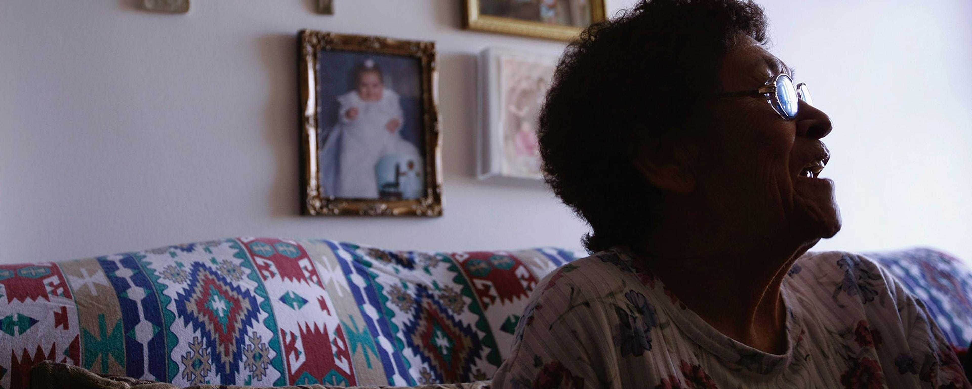 An elderly woman in profile smiling, seated on a patterned sofa with wall art and crosses in the background.