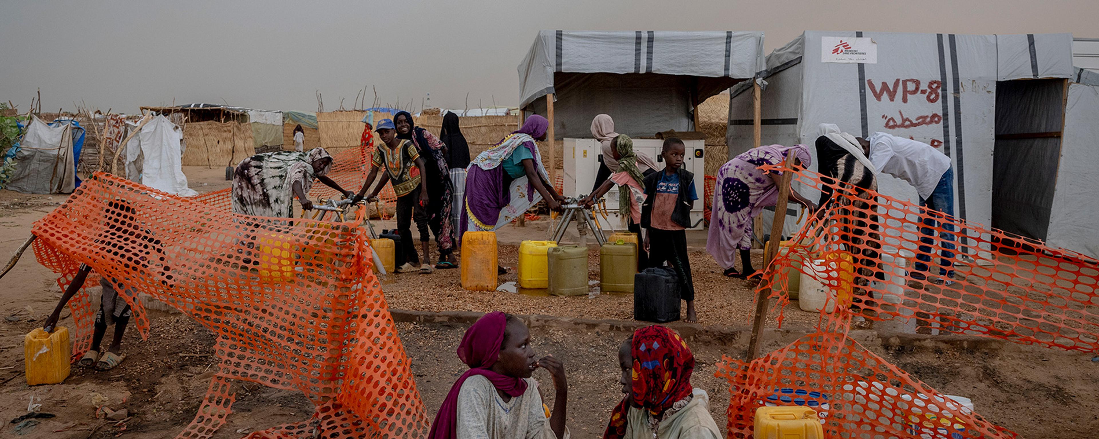 People collecting water from a makeshift station in a camp, surrounded by orange netting and tents in the background.