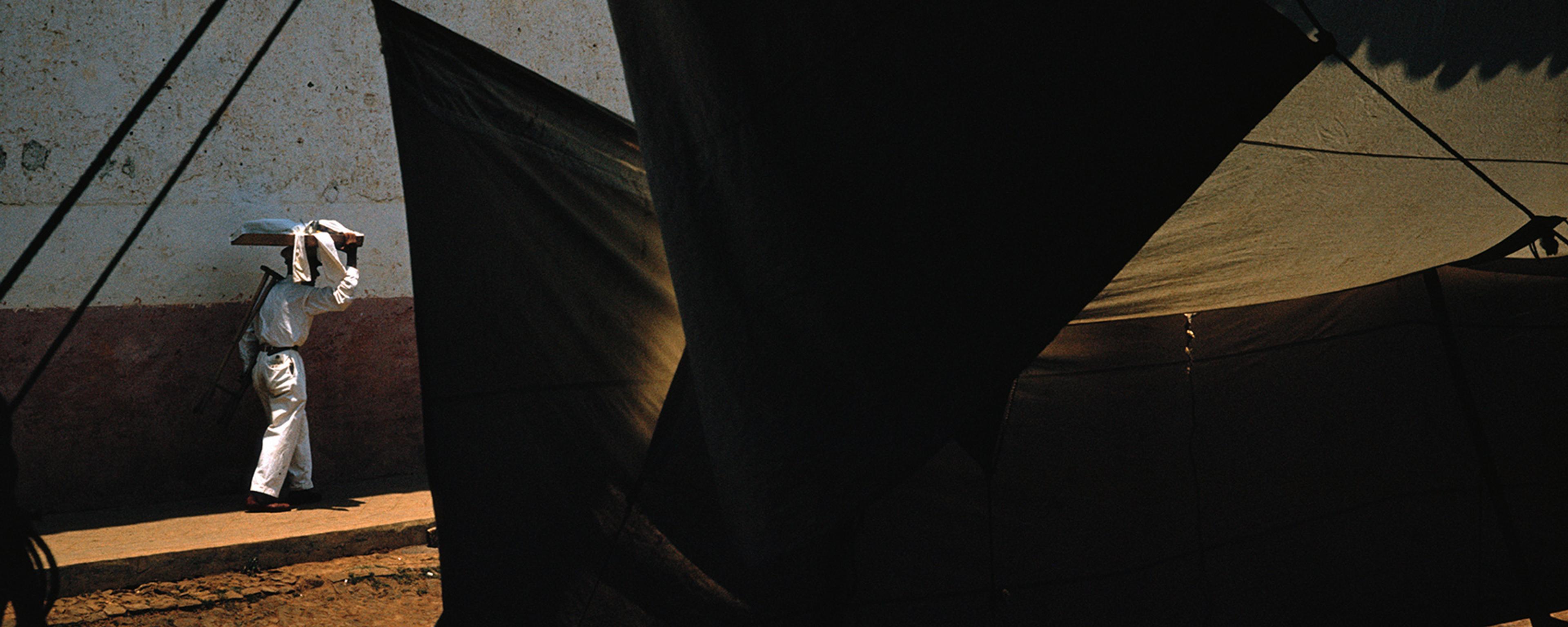 A person in white carrying a load on their head, walking past a dark tent with dramatic light and shadows.