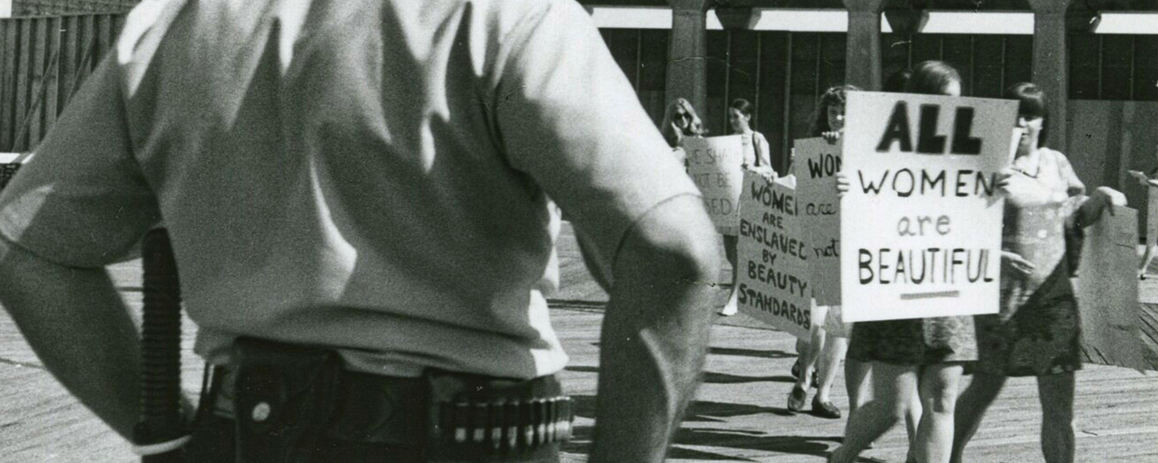 Black and white photo of a policeman observing a women’s protest with signs like “All Women Are Beautiful”