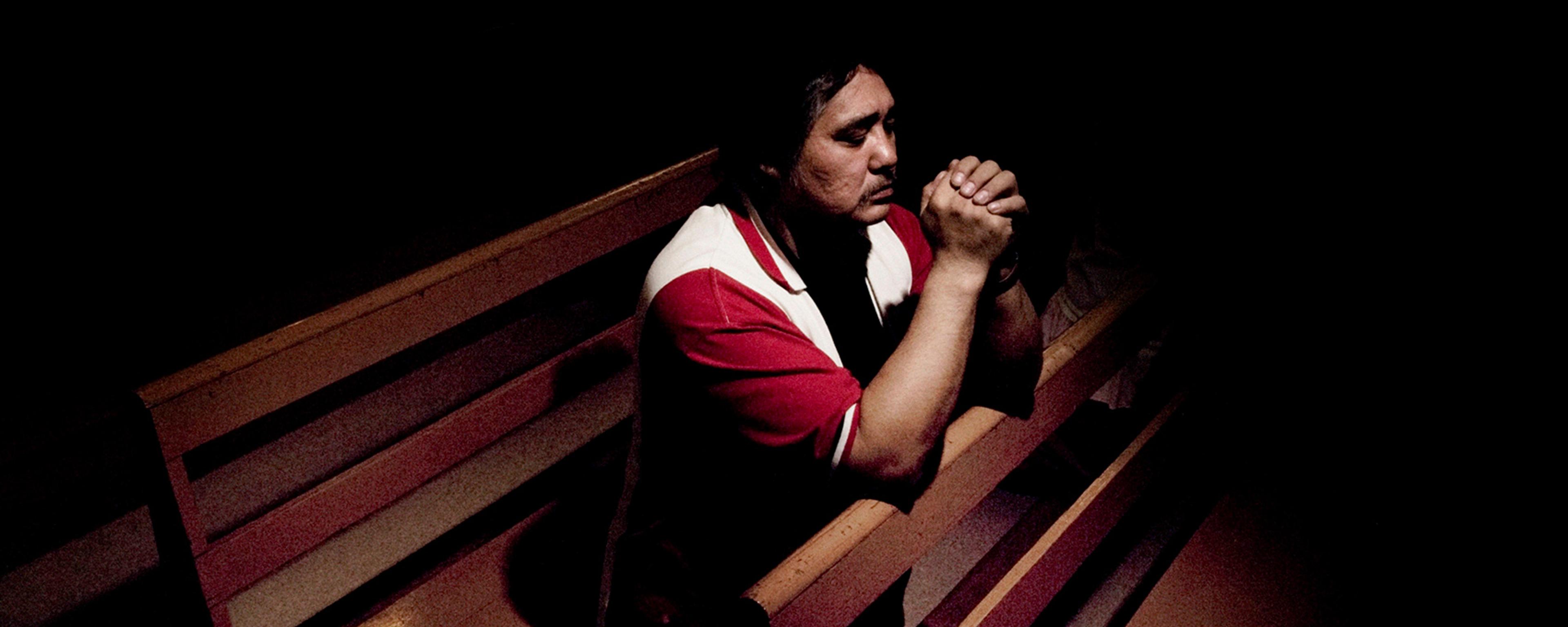 Photo of a man in a red shirt praying on a church pew, hands clasped, with a notebook beside him in dim lighting.