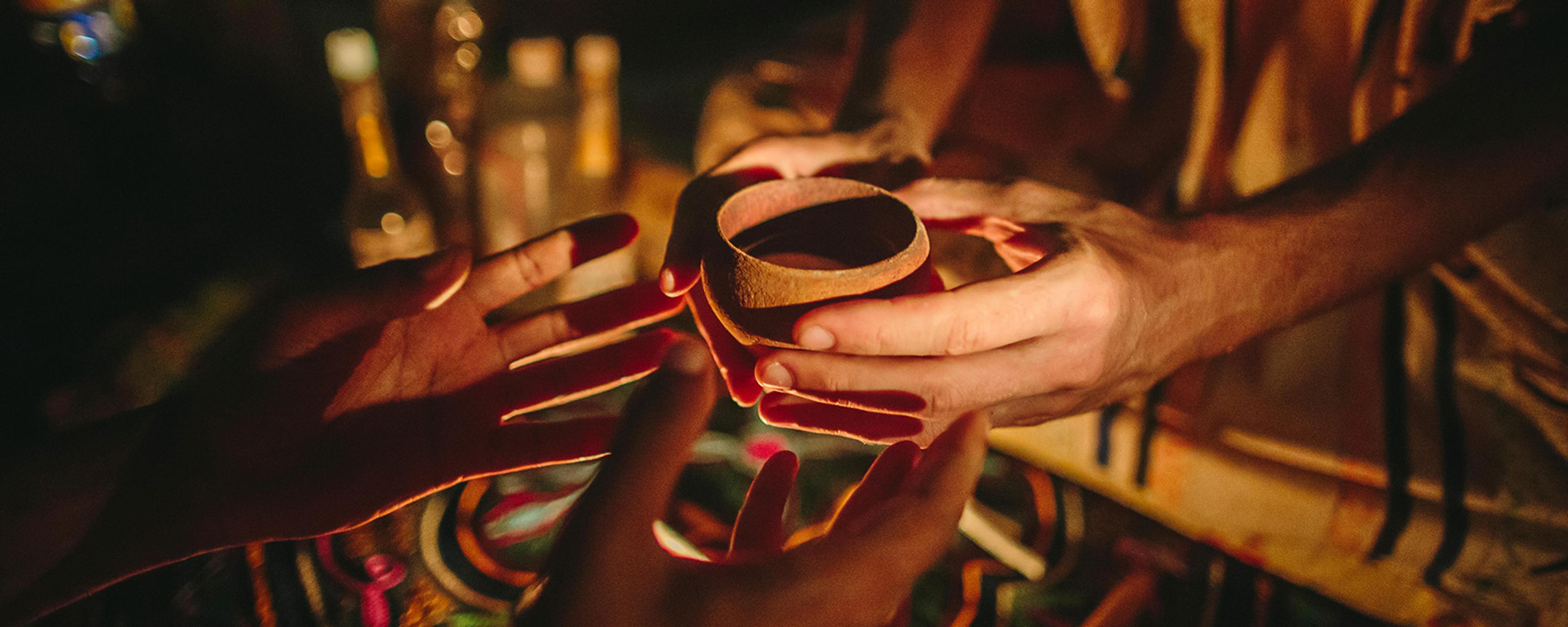 Close-up photo of hands passing a small wooden cup during a dimly lit gathering with bottles visible in the background.