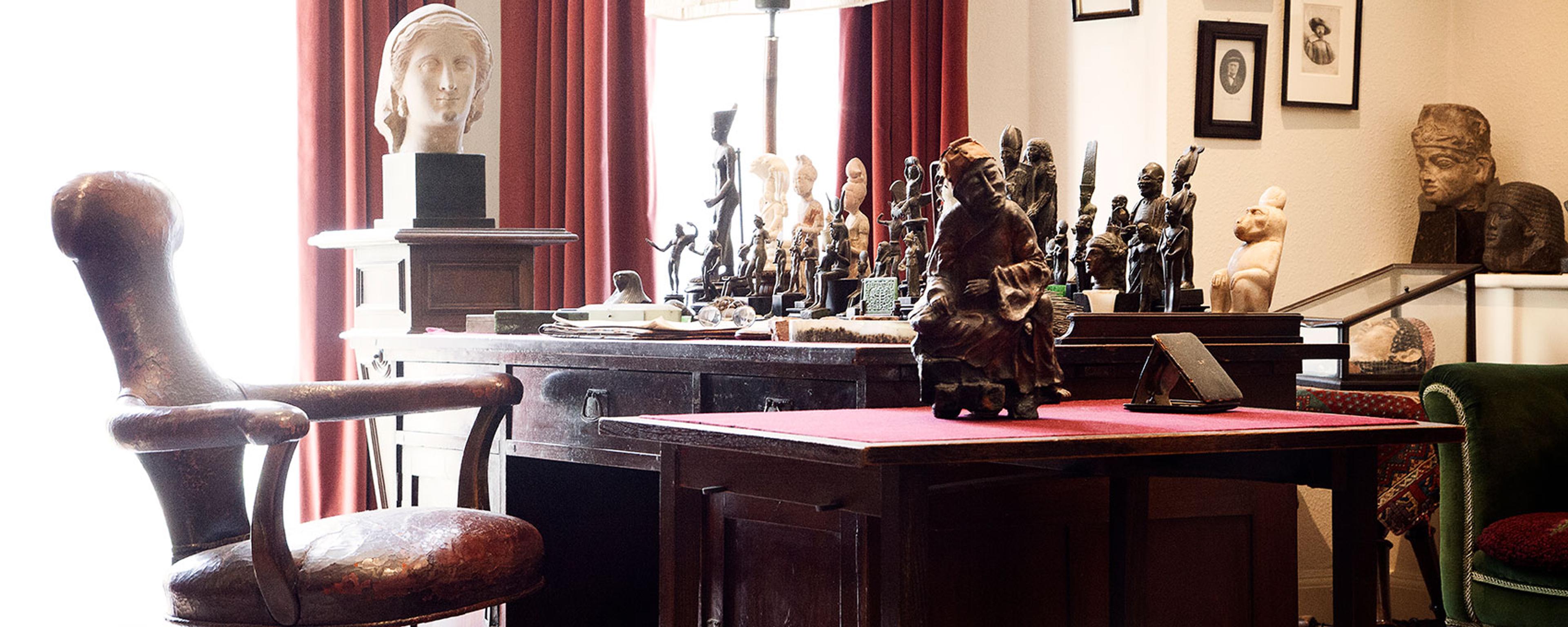 Photo of a study with a wooden desk, red curtains, bust sculptures and a leather chair in a well-lit room.