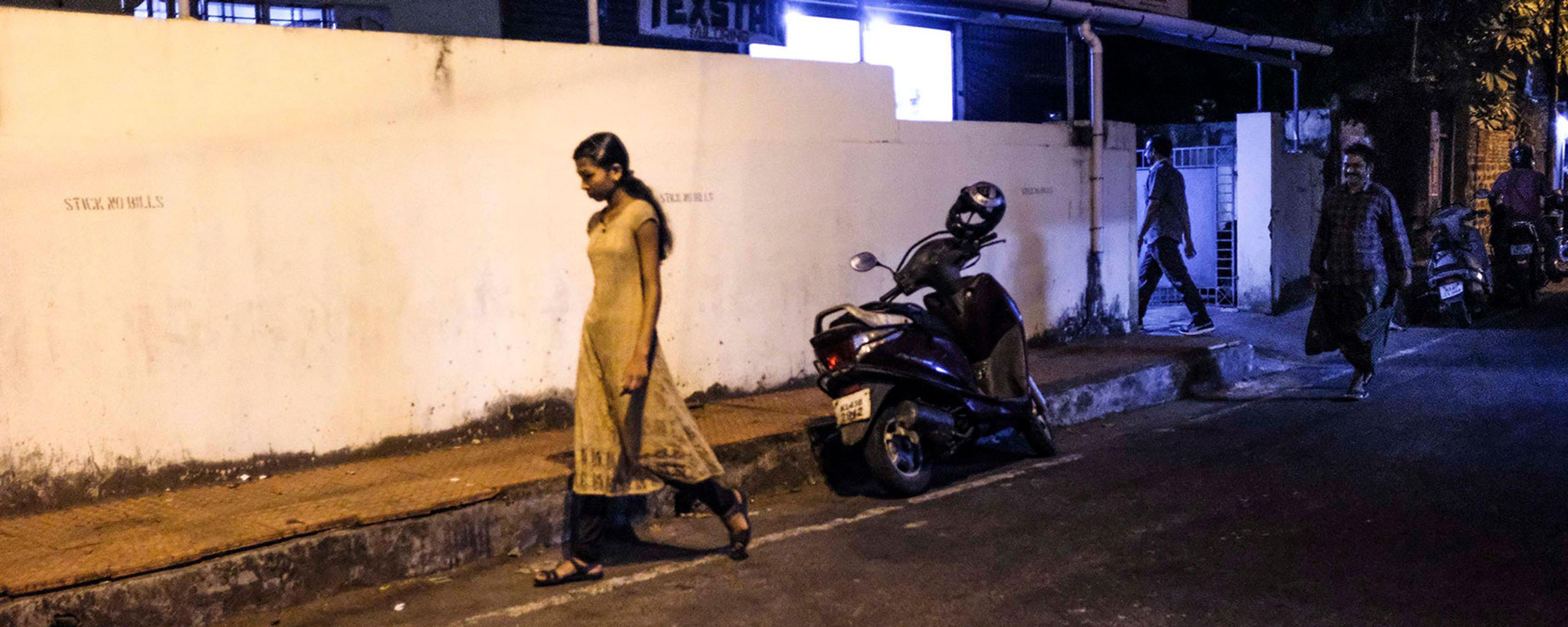 A woman walking on a dimly lit street pavement at night with a parked scooter and two men walking in the background, one appearing to follow her; the woman looks downcast, the man appears to be smiling.