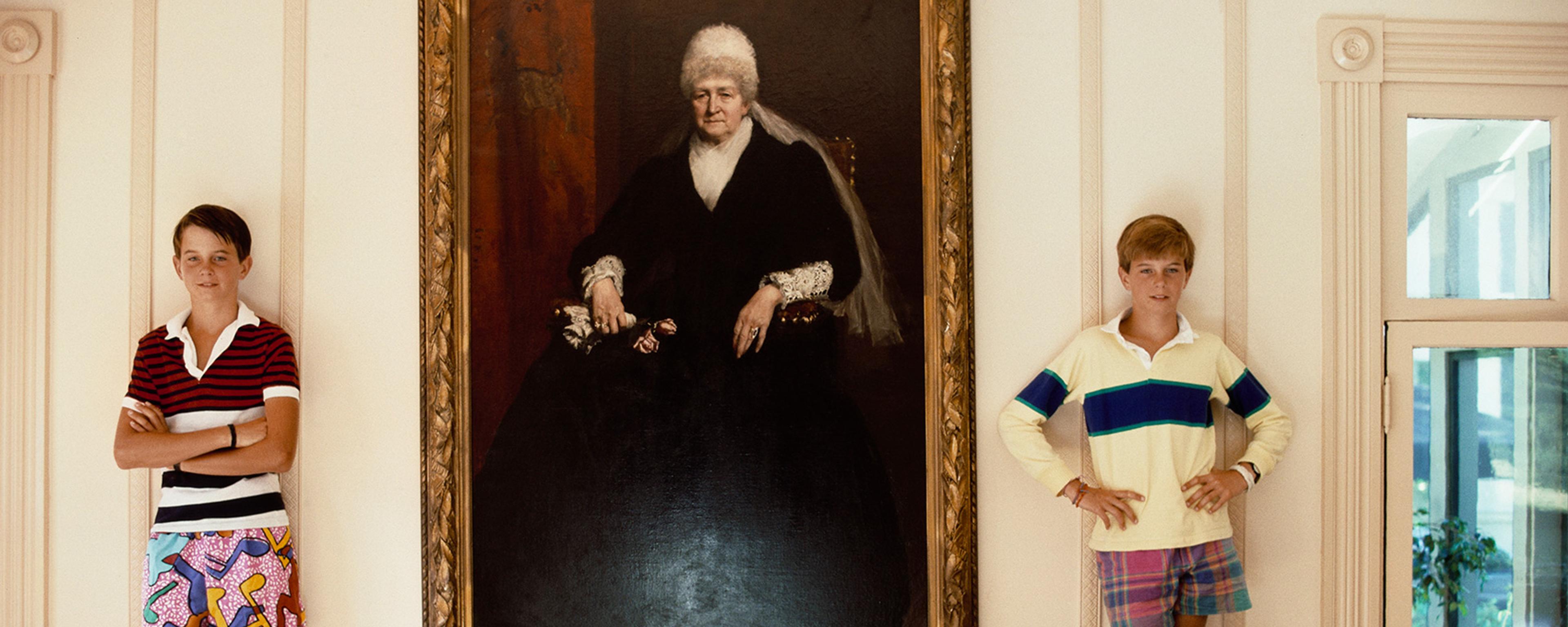 Two boys in colourful clothes standing beside a large painting of an elderly woman in a gold frame.