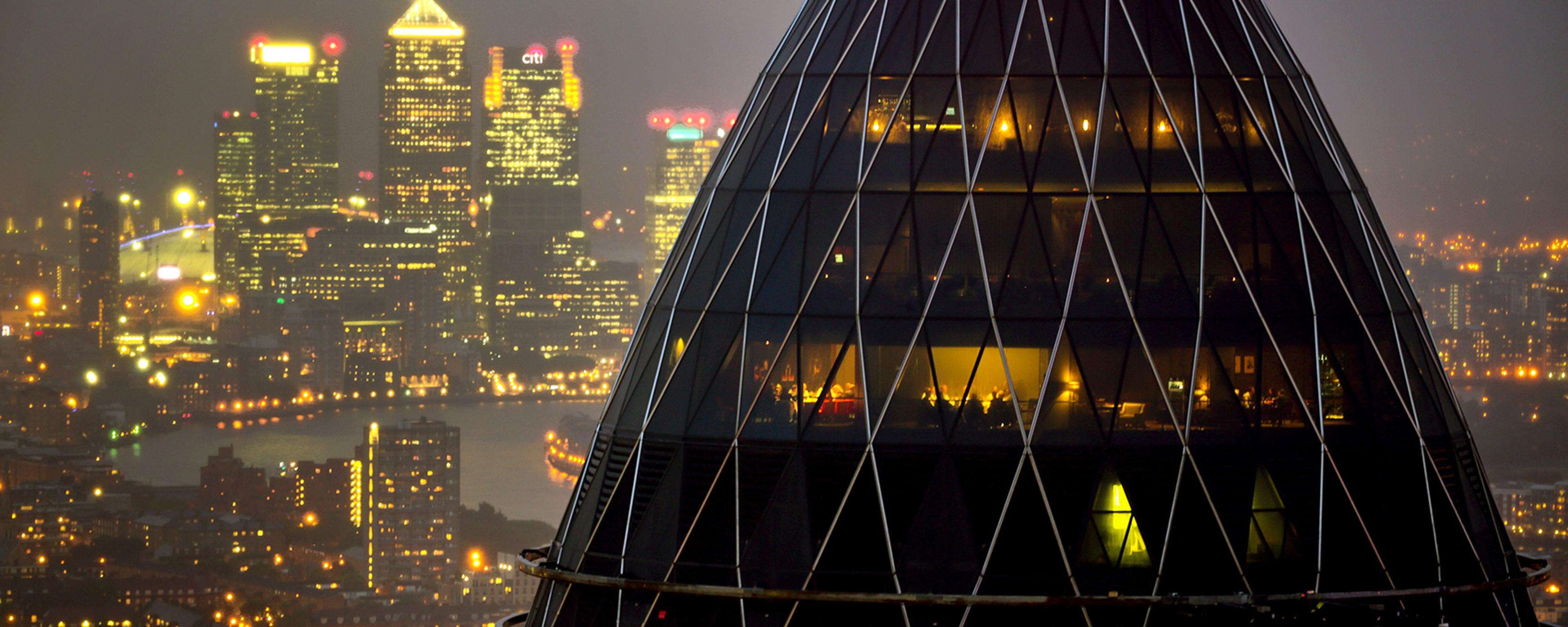 London’s Gherkin building lit at night with Canary Wharf skyscrapers glowing in the hazy background.