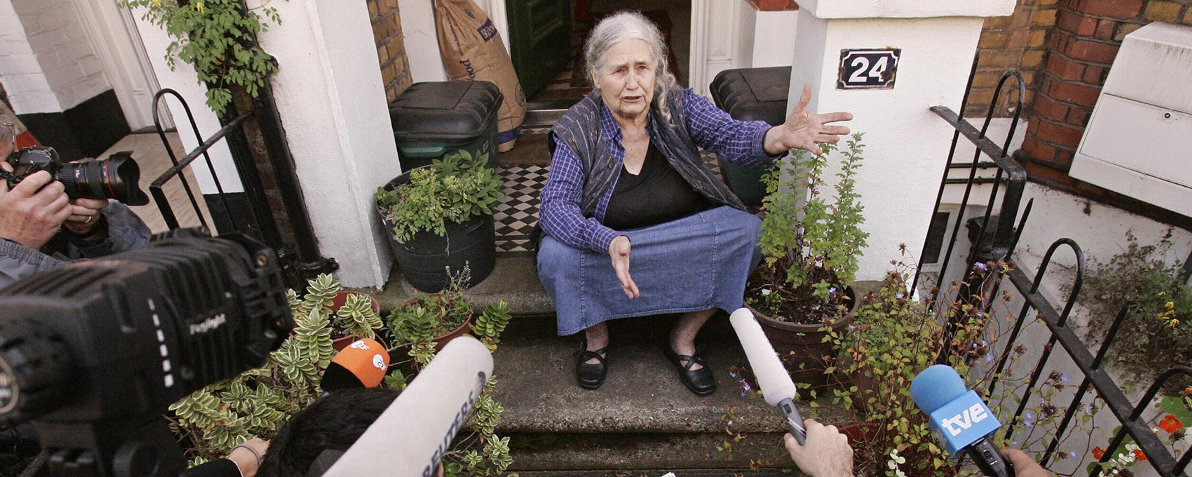 An older woman sitting on steps, speaking to journalists holding microphones and cameras outside a house.