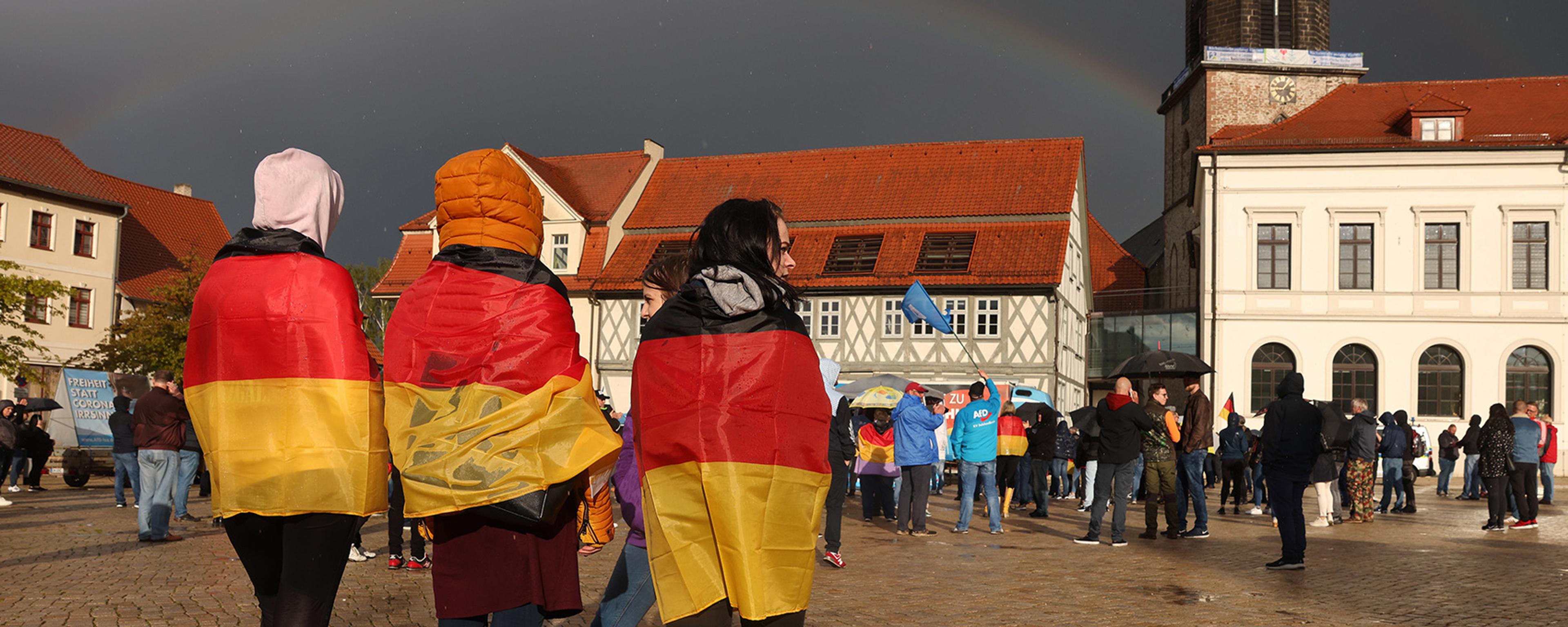 Three people draped in German flags standing in a town square during a gathering, with a rainbow in the cloudy sky.