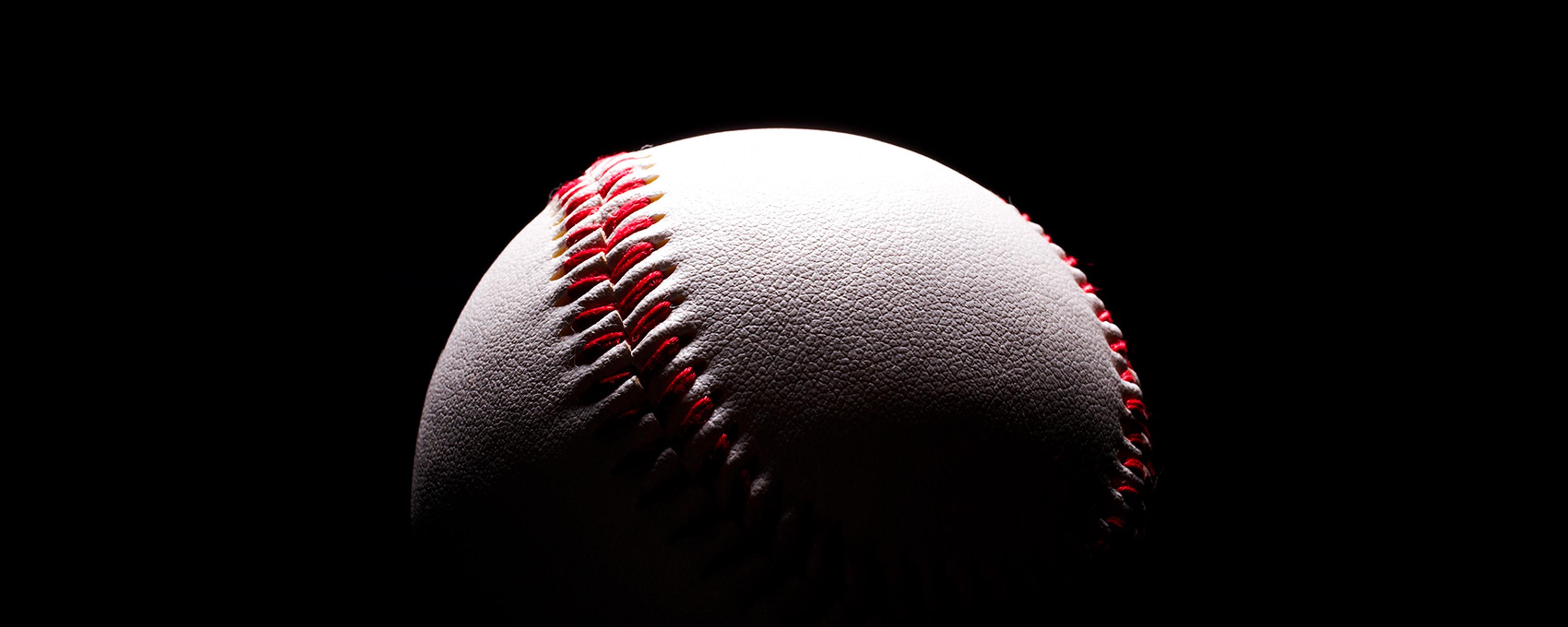 A baseball with red stitching on a black background partially lit from the top left corner.