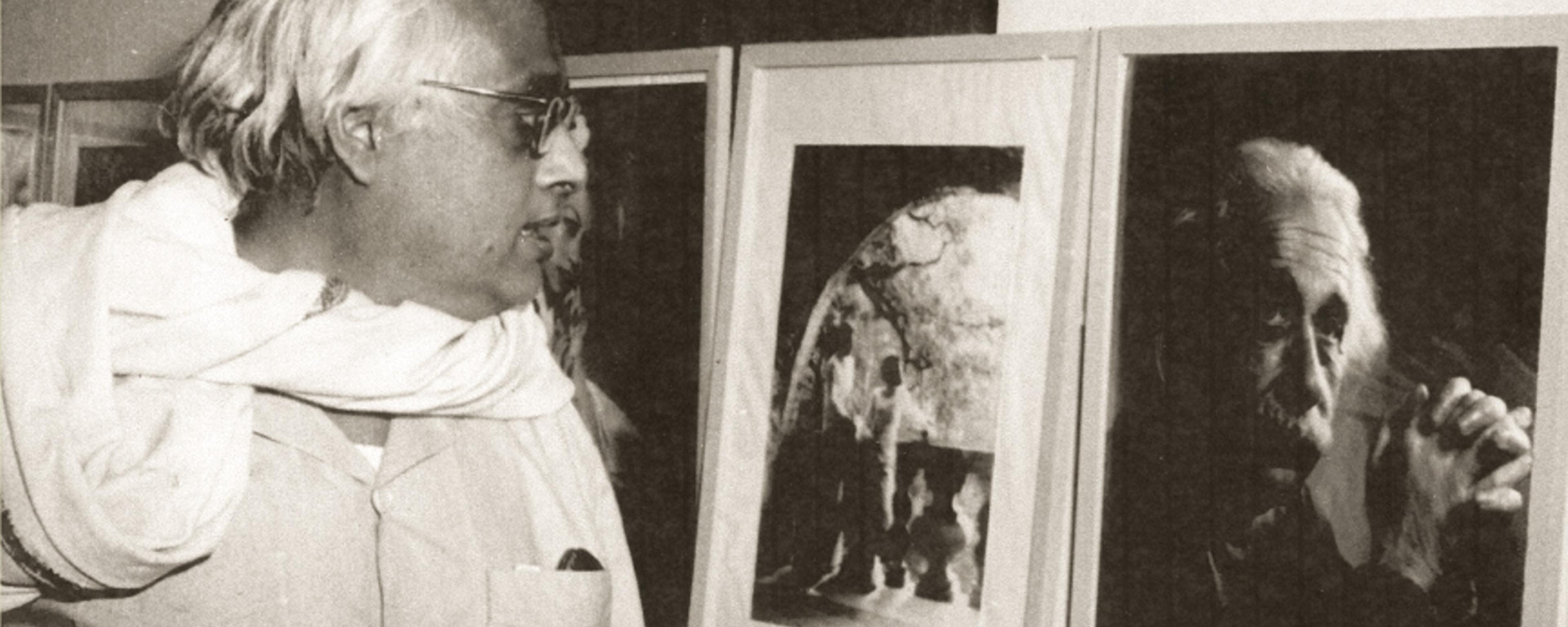 Black-and-white photo of a man viewing framed artworks on a wall in an exhibition setting.