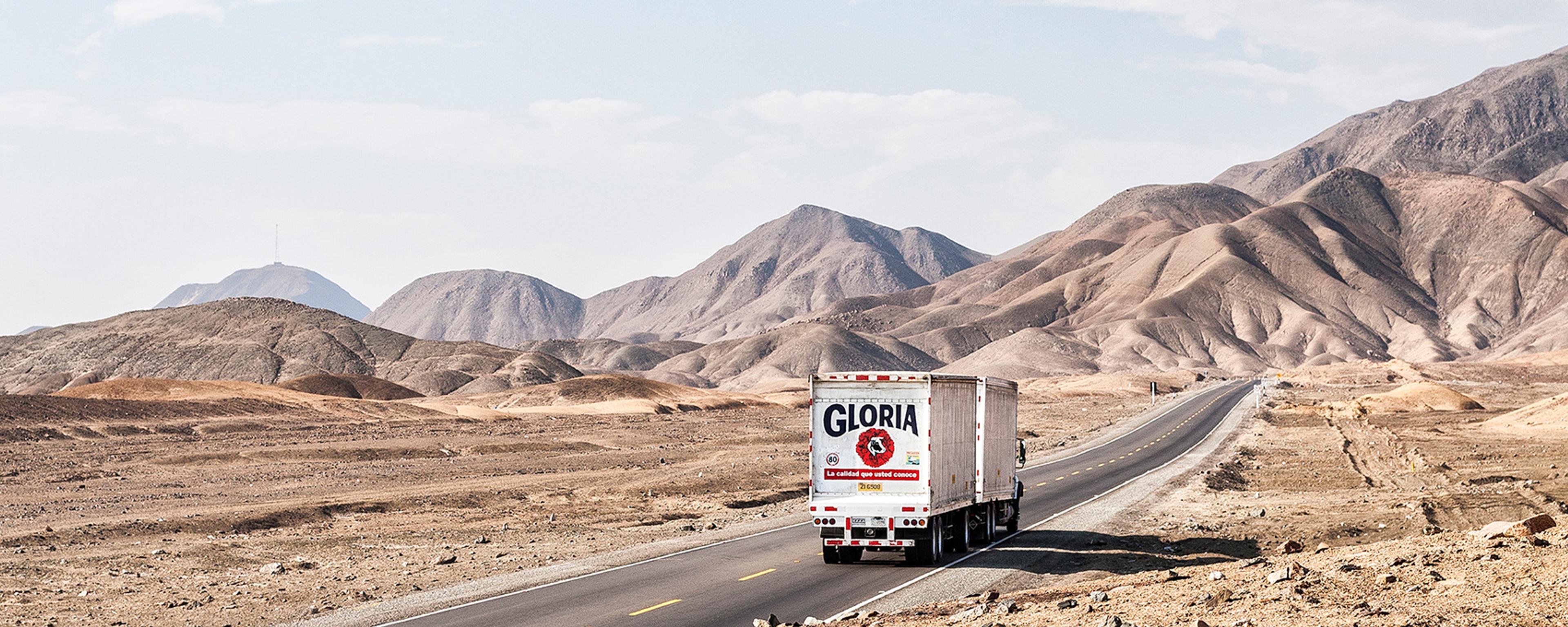 A lorry on a long desert road with rocky terrain and mountains in the background under a partly cloudy sky.