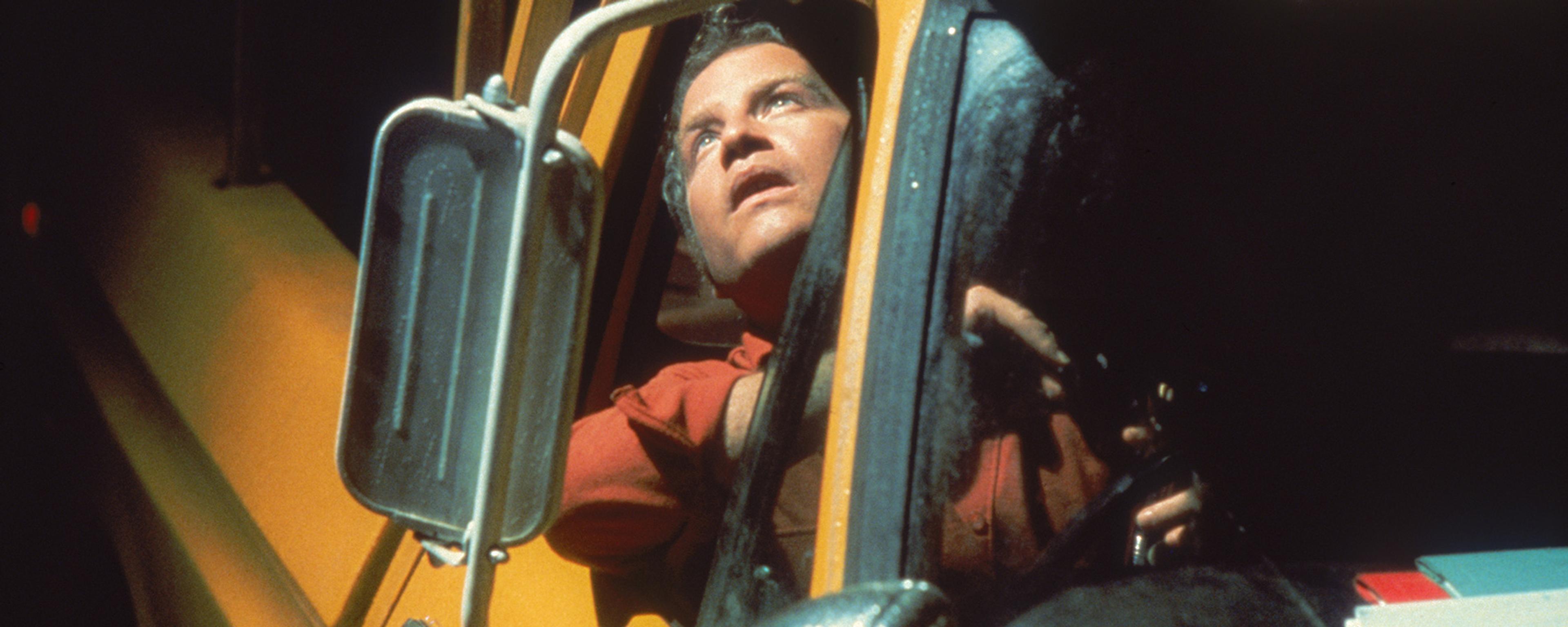 A man looking up from the driver’s seat of a yellow vehicle with an open window and side mirror visible.
