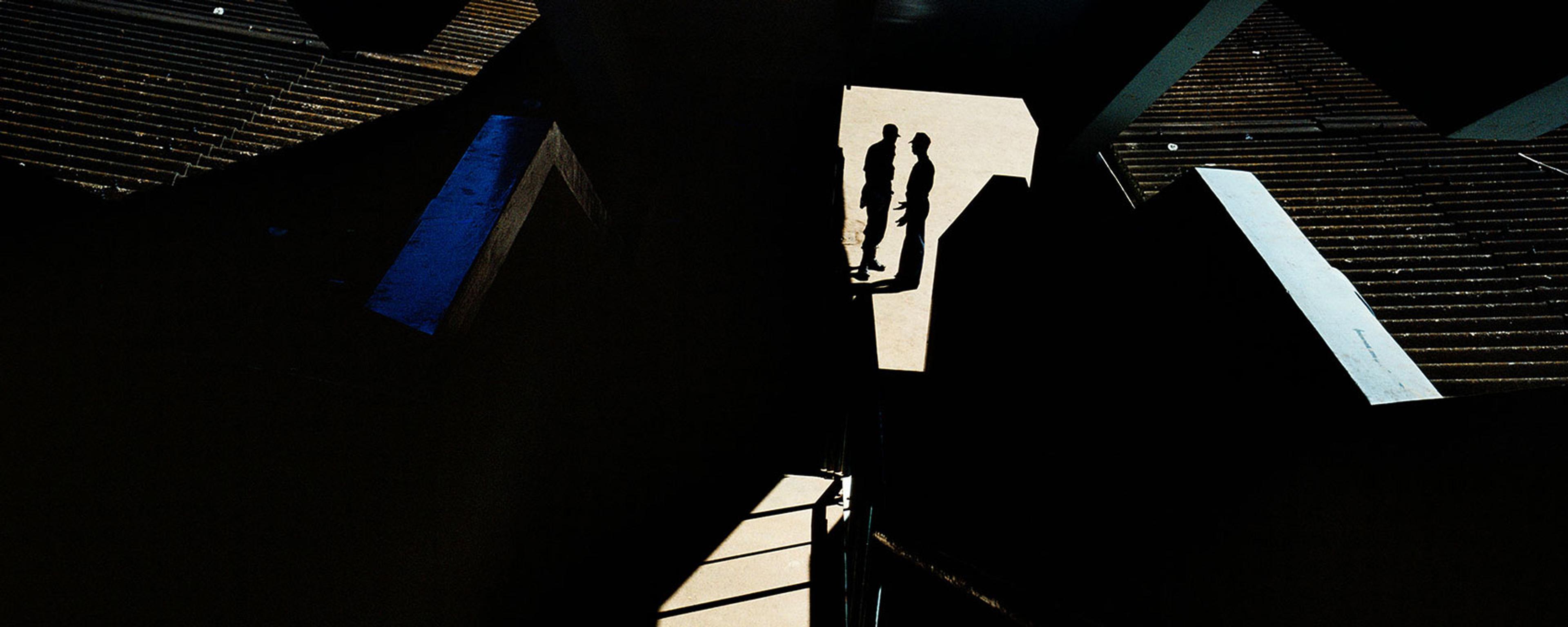 Photograph of two silhouetted figures on a sunlit path surrounded by dark geometric shadows and structures.