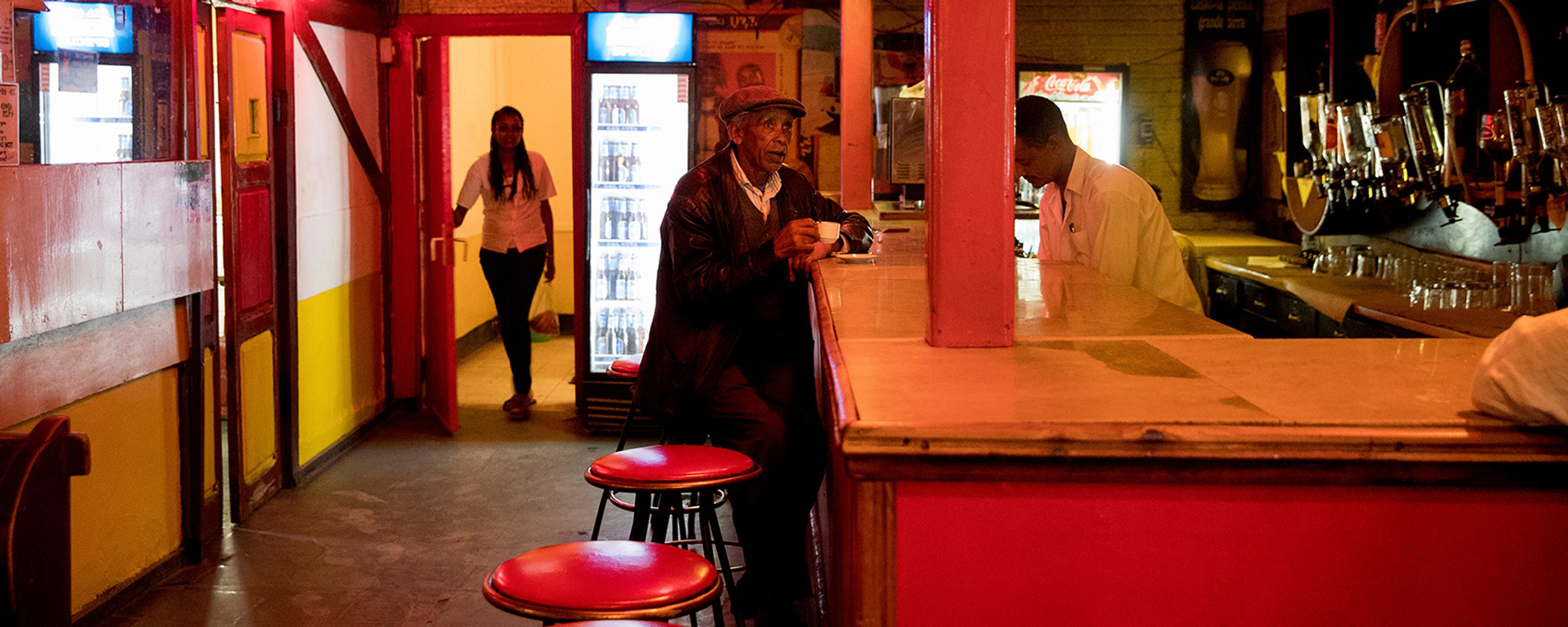 A dimly lit bar with red decor, a man sitting at the counter, a bartender, and a woman entering through a door.