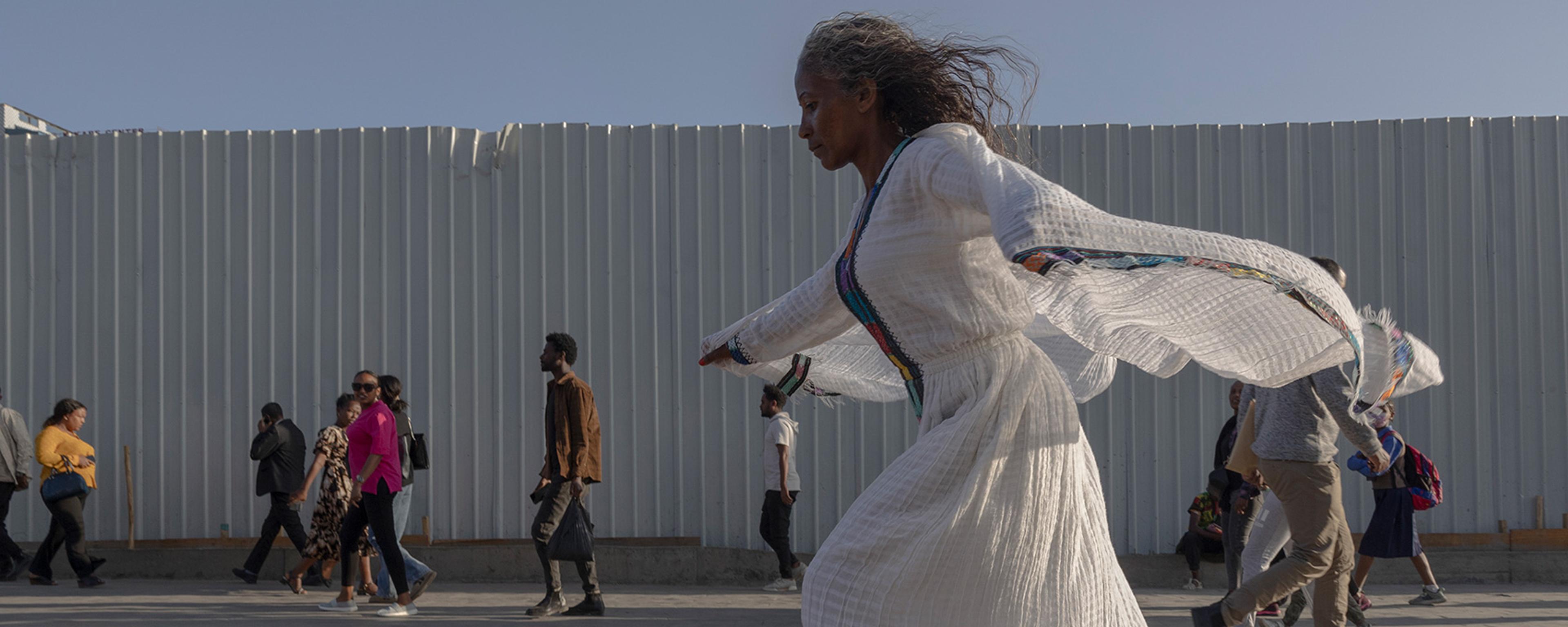 A woman in a flowing white dress skateboarding past people walking on a sunny day next to a corrugated fence.