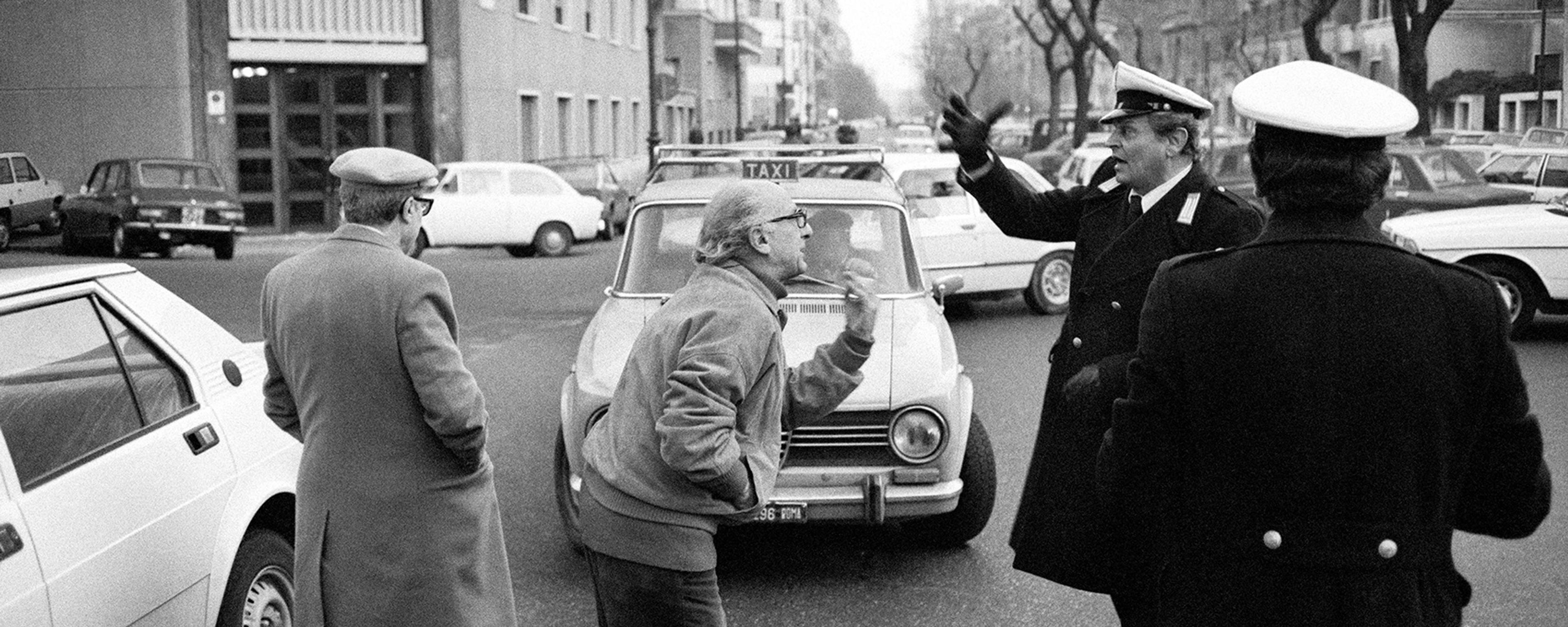 Black and white photo of a street scene with a heated discussion involving a traffic officer and men near cars.