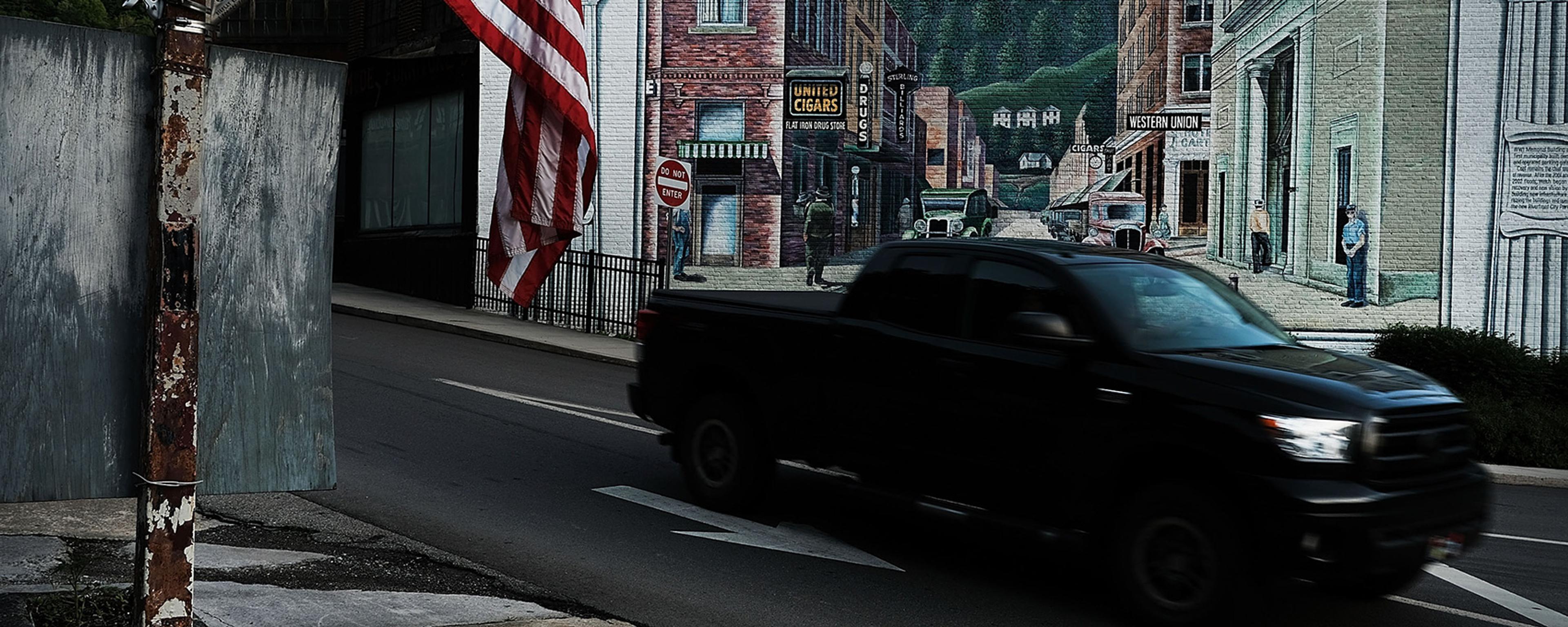 A street with an American flag a mural of a vintage town scene and a black pickup truck passing by.