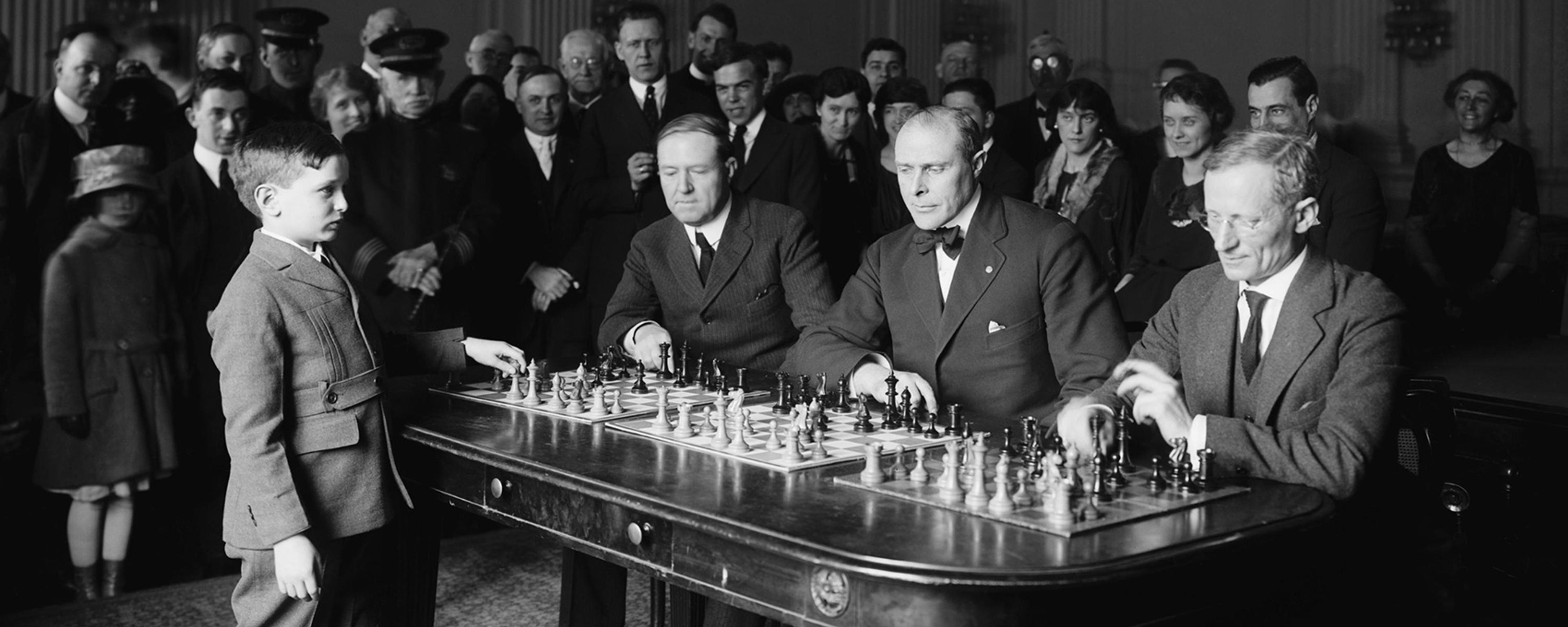 Black and white photo of a young boy playing simultaneous chess against three suited men watched by a crowd.