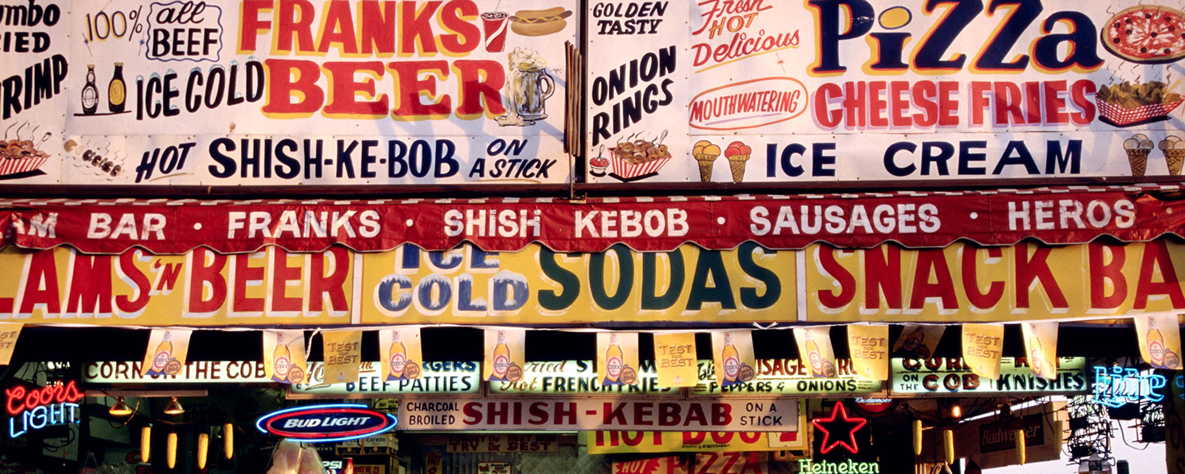 Photo of a vibrant snack bar with colourful signs advertising beer, pizza, sausages and ice cream.