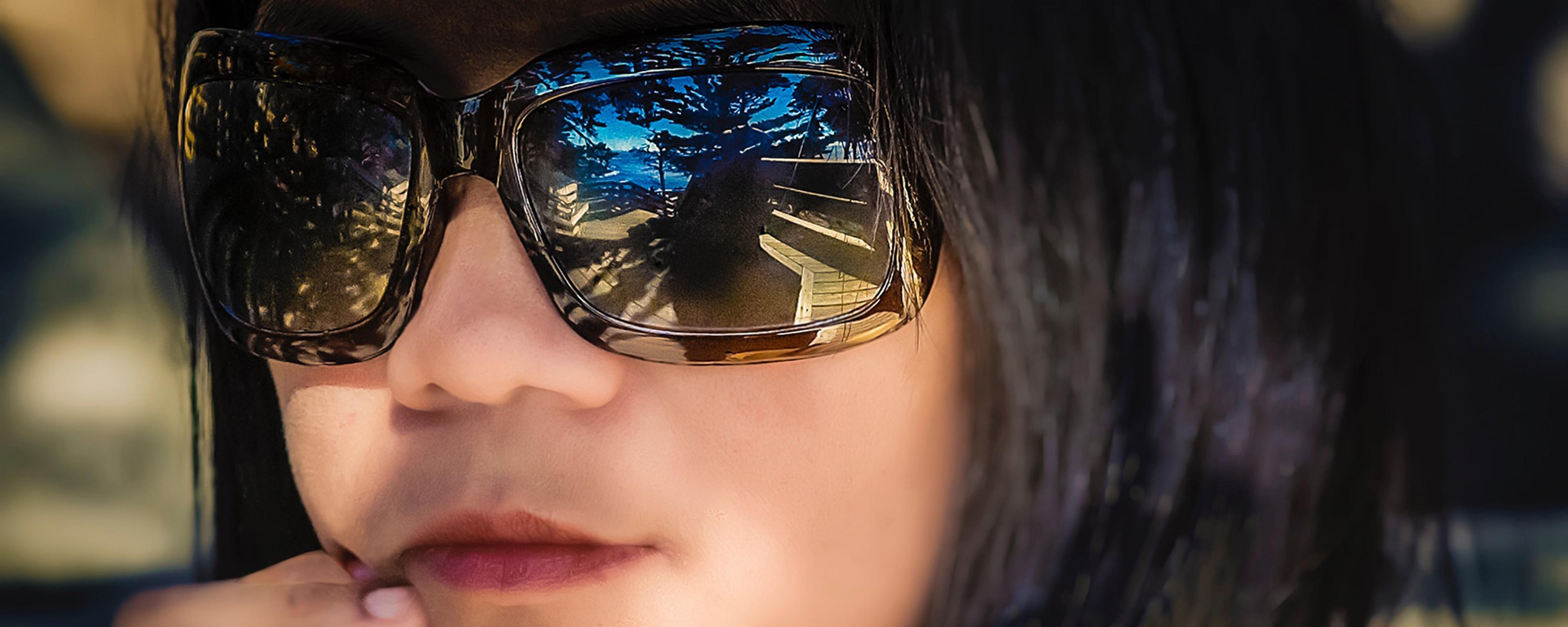 Photo of a person wearing large sunglasses with outdoor scenery reflected, resting their fingers on their chin in a thoughtful pose.