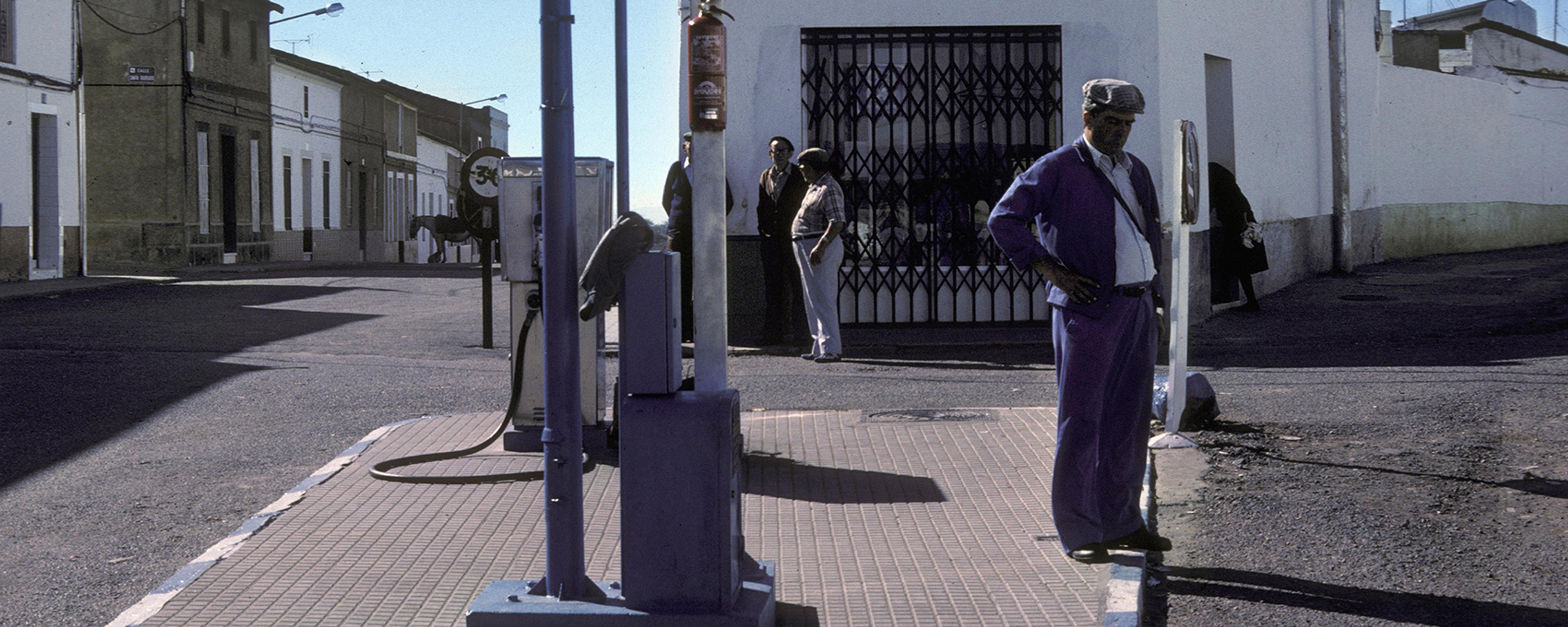 A man standing near a petrol pump on a traffic island between two diverging roads under a clear blue sky.