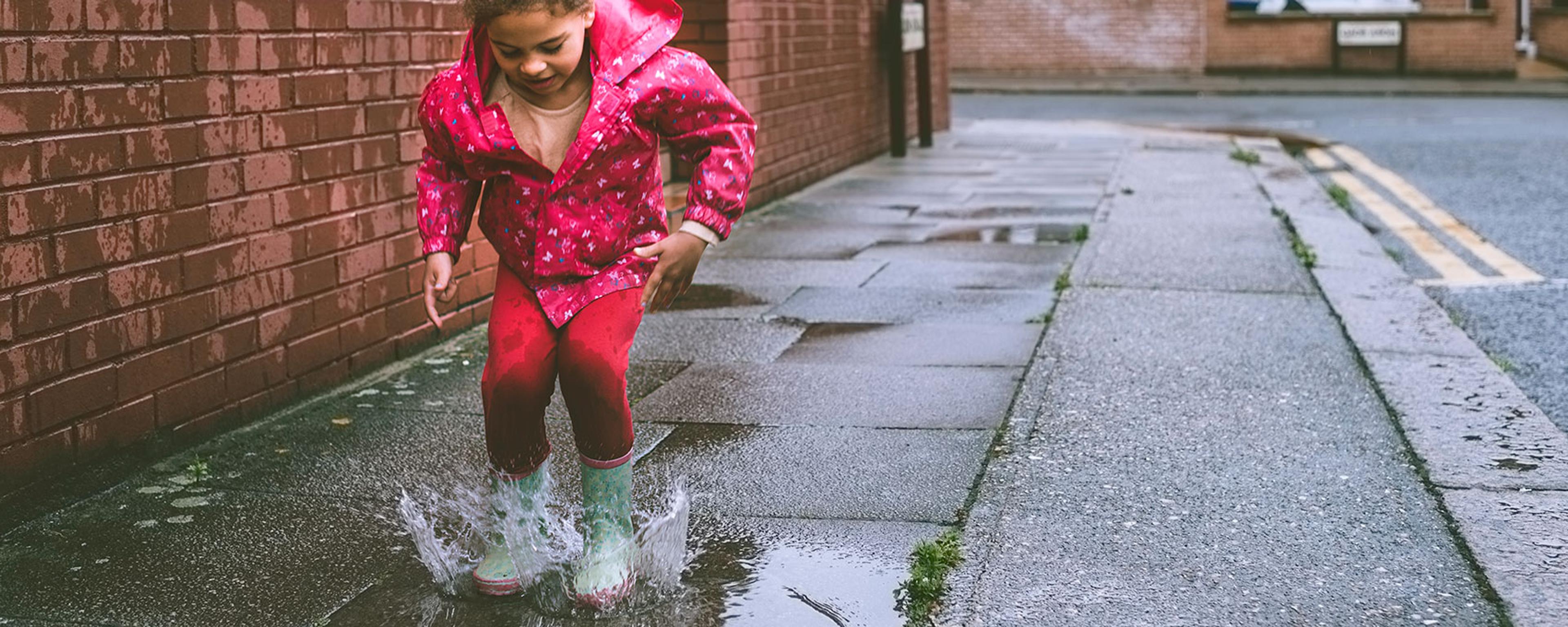 A child in a pink raincoat jumping in a puddle on a city street on a rainy day with terraced houses in the background.