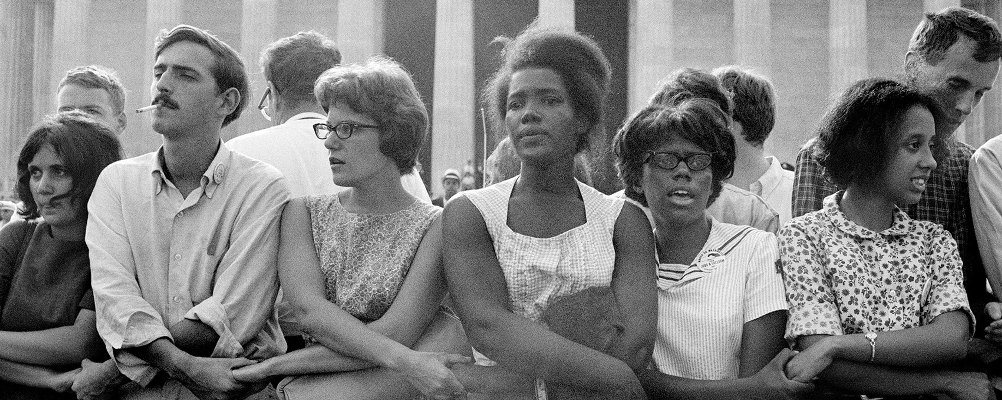 Black and white photo of a diverse group of people standing arm in arm in front of a columned building, showing solidarity.