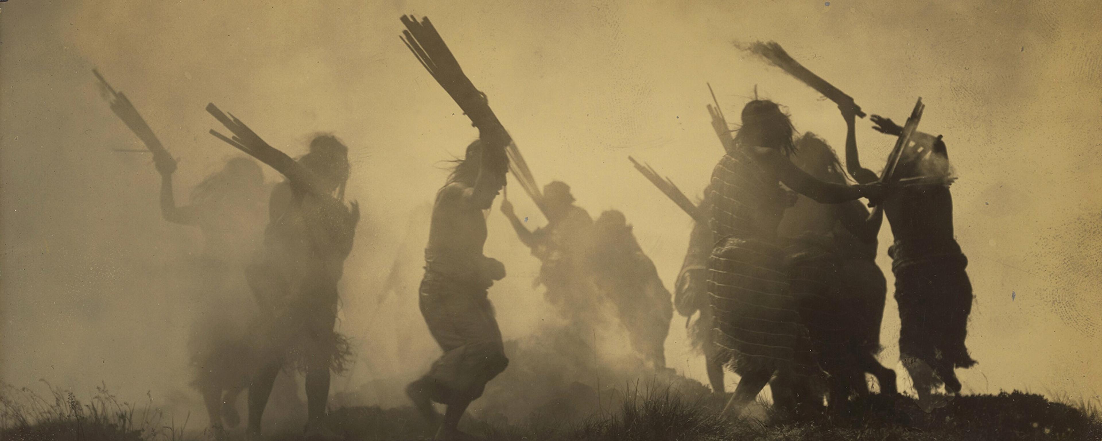 Sepia-toned photo of silhouetted figures dancing with sticks in a smoky environment, creating a dramatic atmosphere.