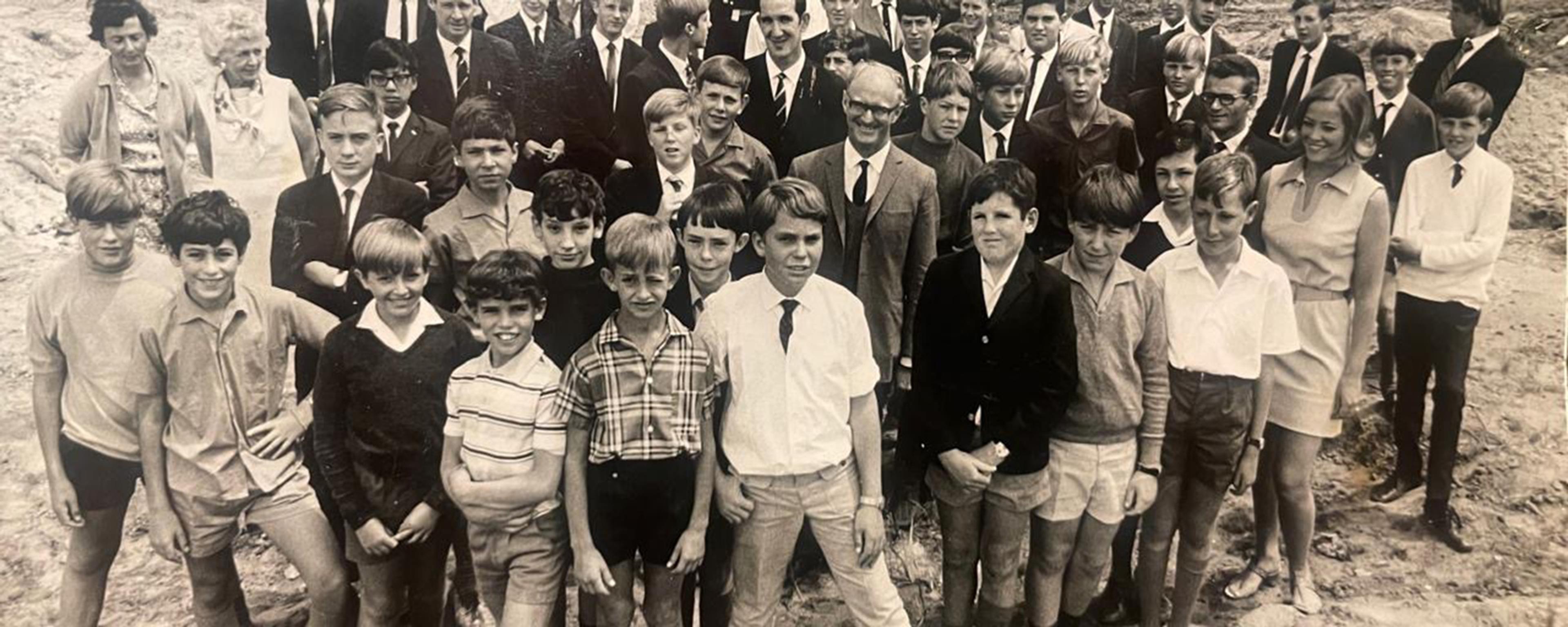 Black and white photo of a large group of boys and a few adults standing on bare ground, all dressed in 1960s attire.