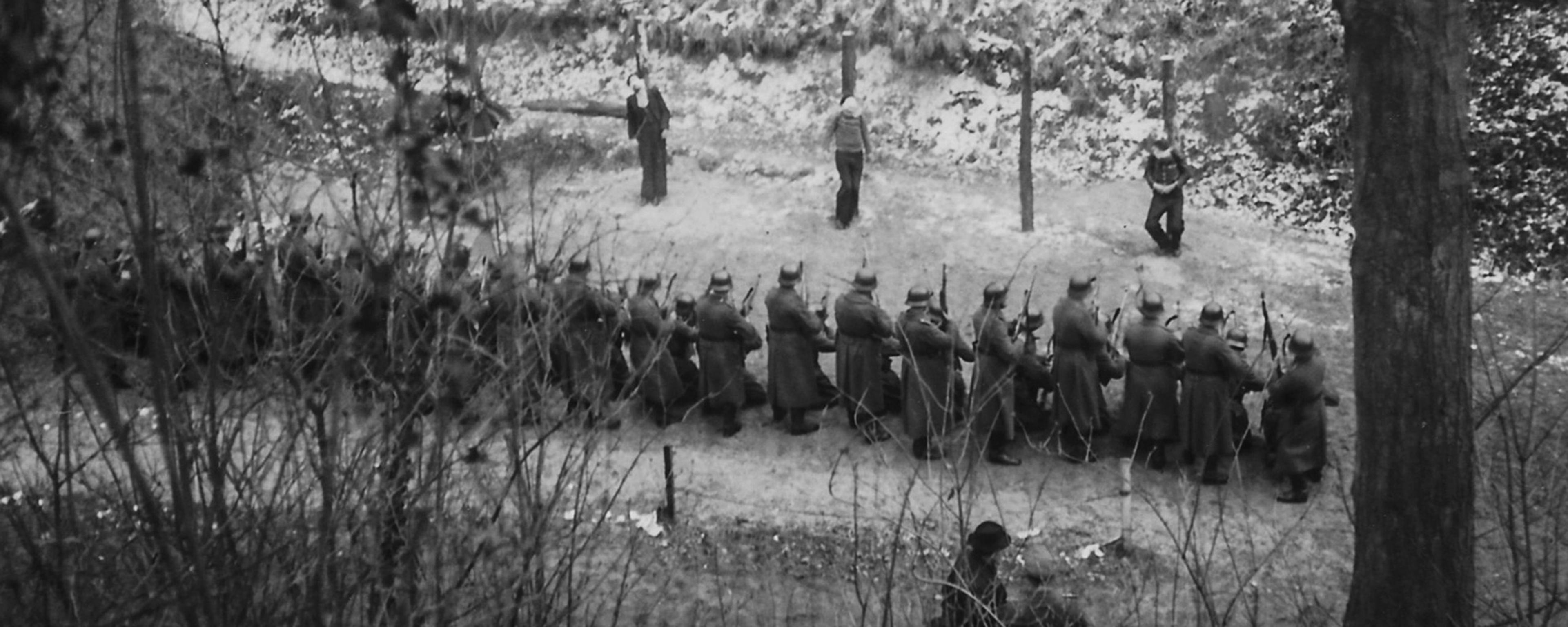 Black and white photo of a firing squad in a wooded area with snow on the ground, soldiers aiming rifles at standing figures.