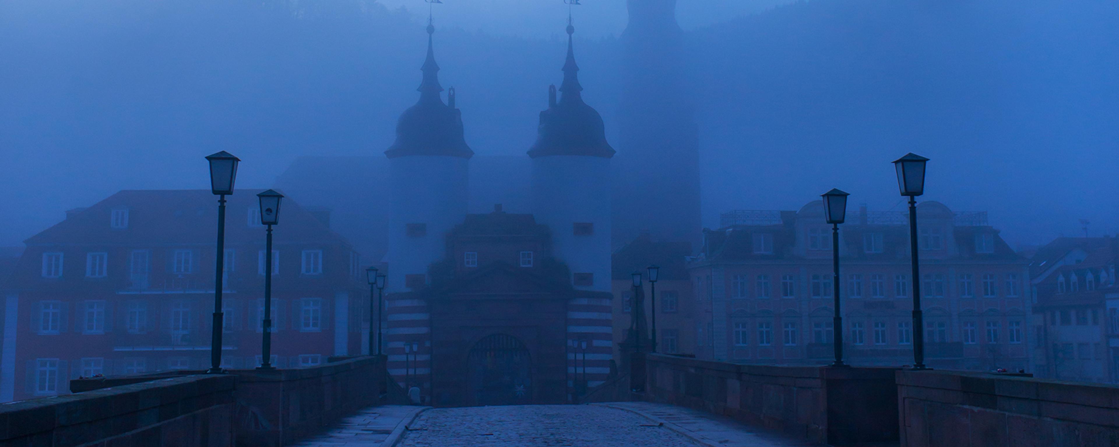 An early morning view across an old bridge towards the spires of a historic medieval city partially obscured by fog
