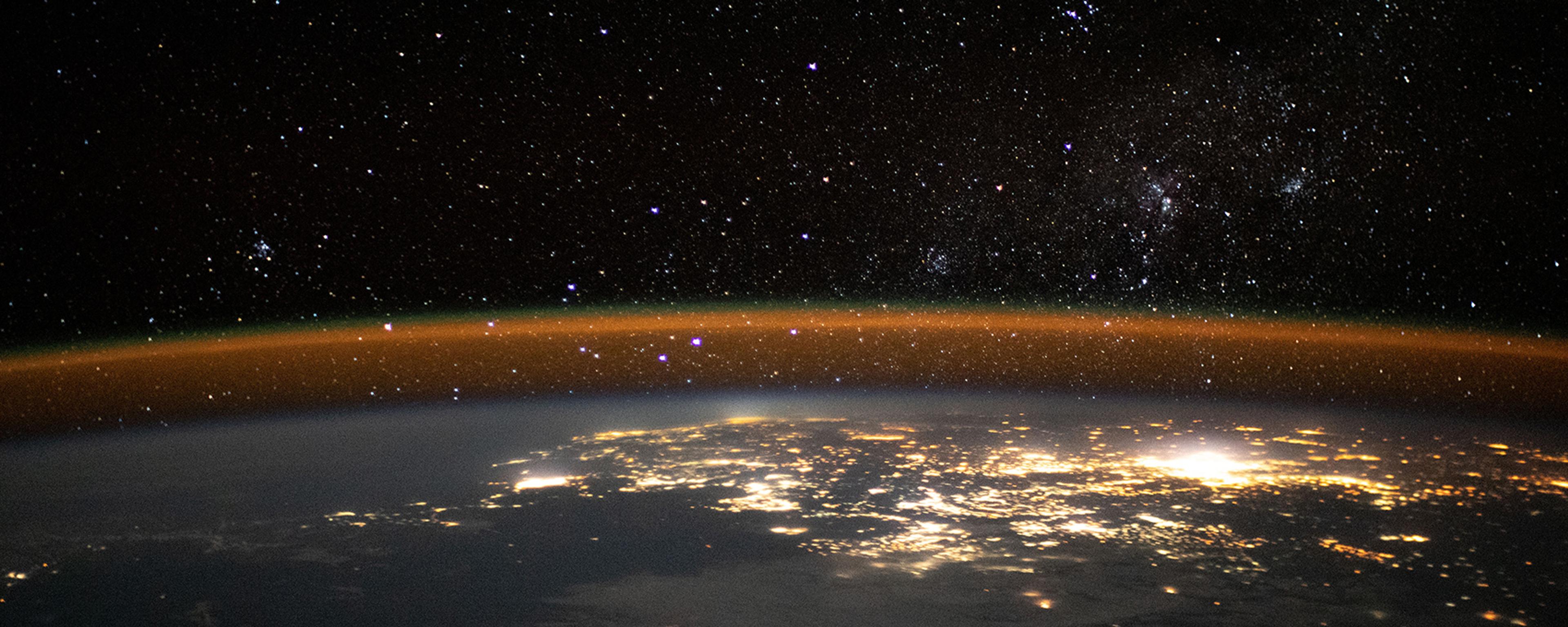 An image shows the earth horizon at night seen from space. The lights of a city glow beneath the vast starry night of space
