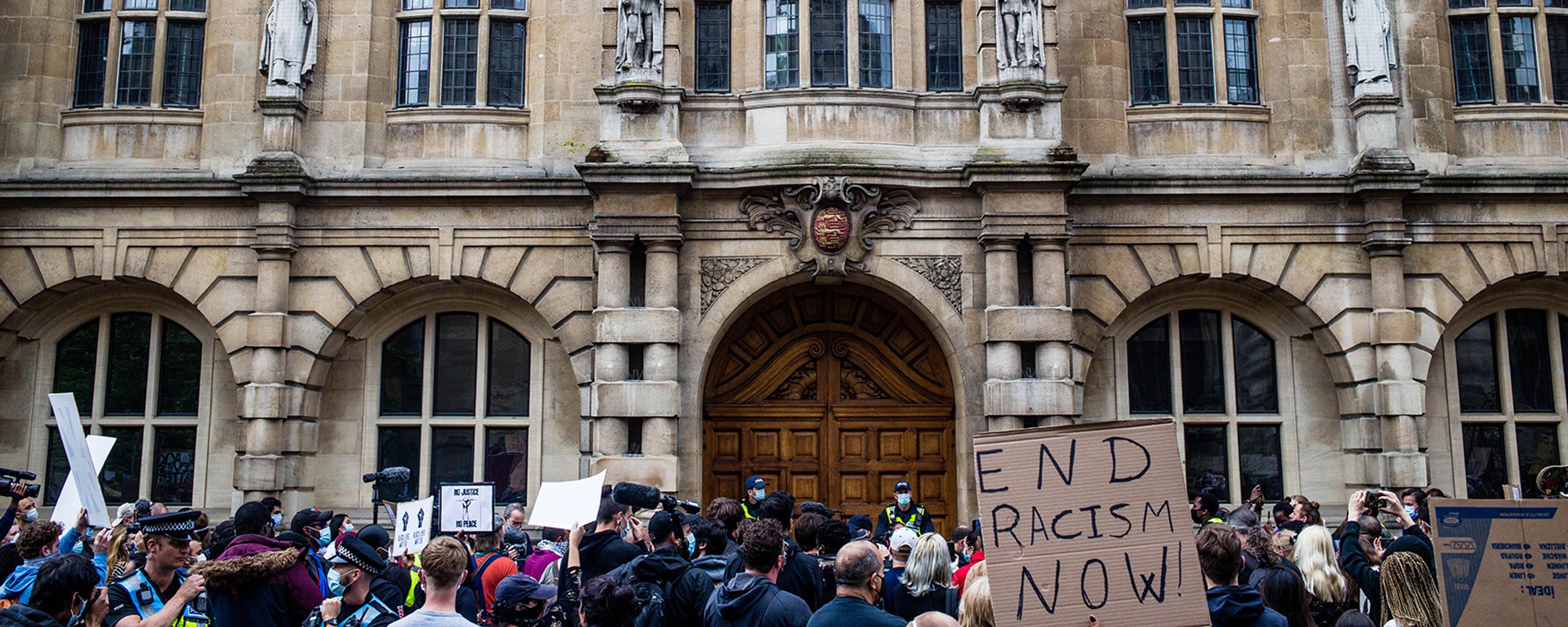 Photo of a protest in front of a historic building with statues. Signs read “End Racism Now!” and other messages.