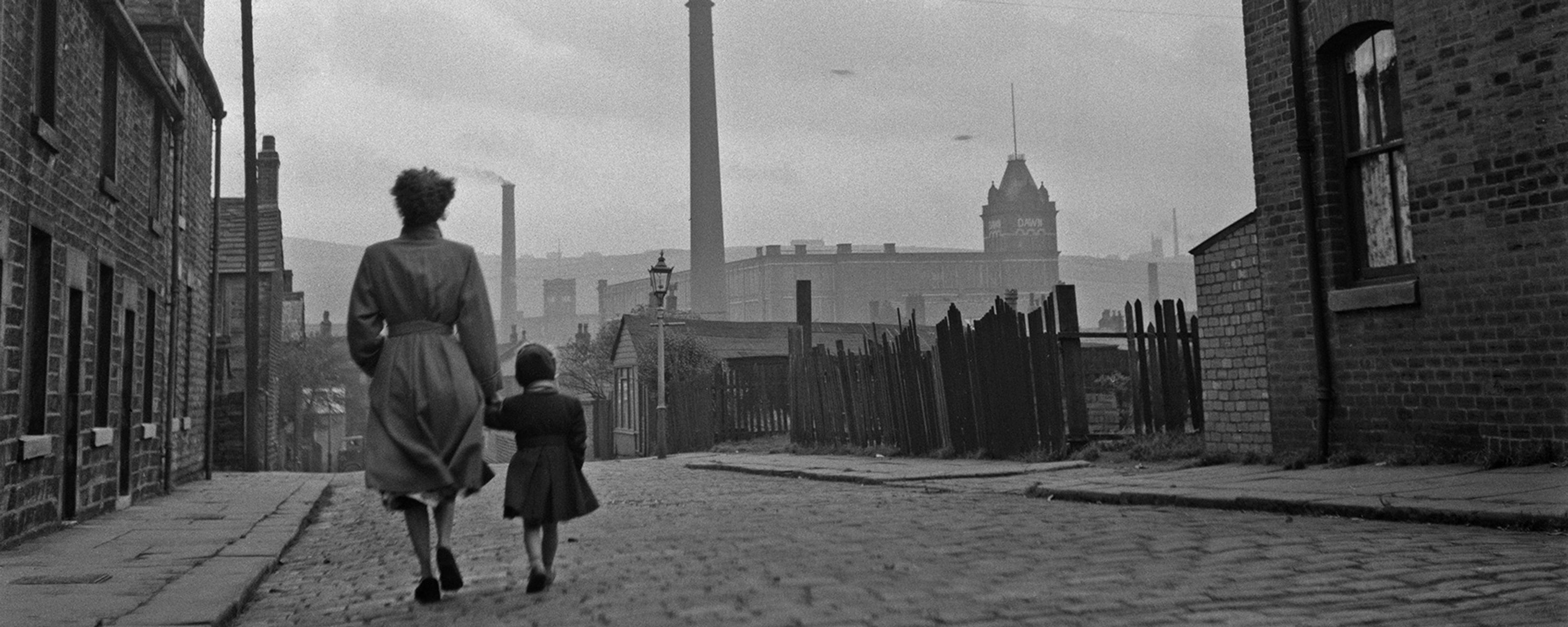 Black and white photo of a woman and child walking on a cobbled street in an industrial town with chimneys in the background.