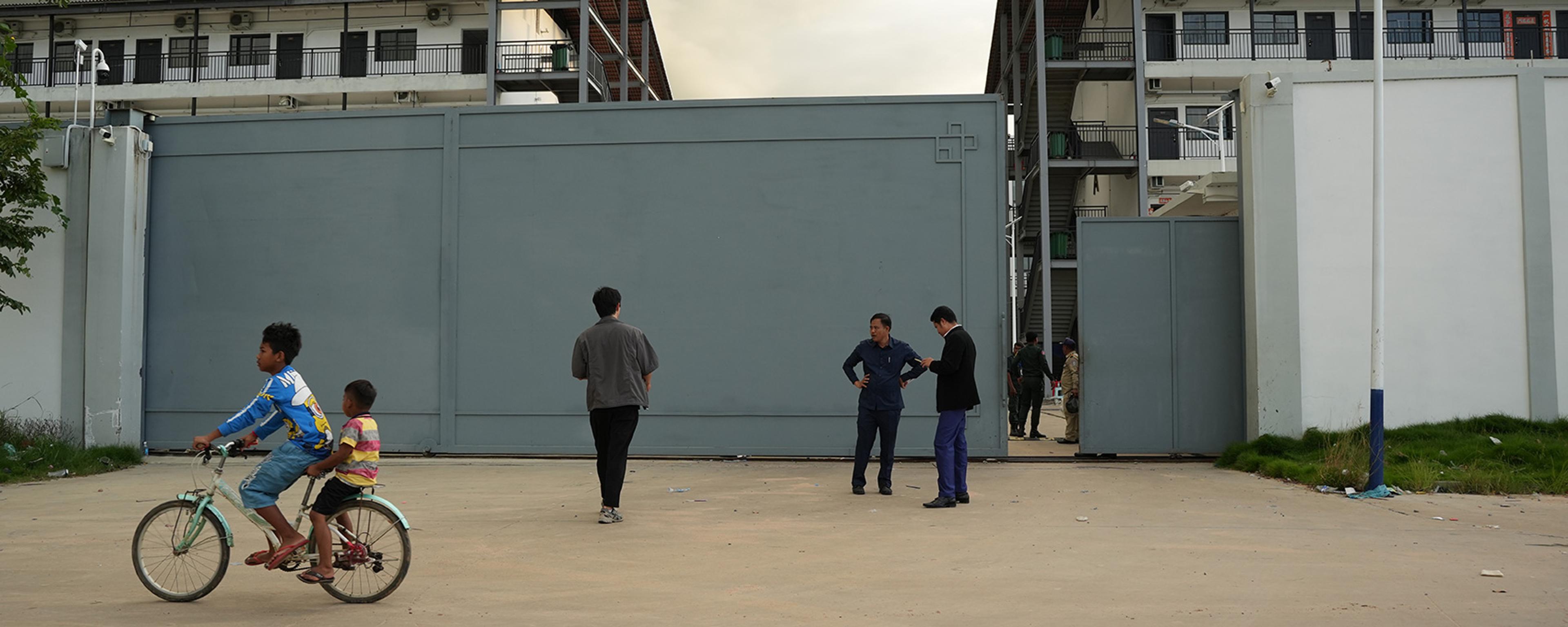 Two children on a bike passing a group of people standing outside a large metal gate with buildings in the background.