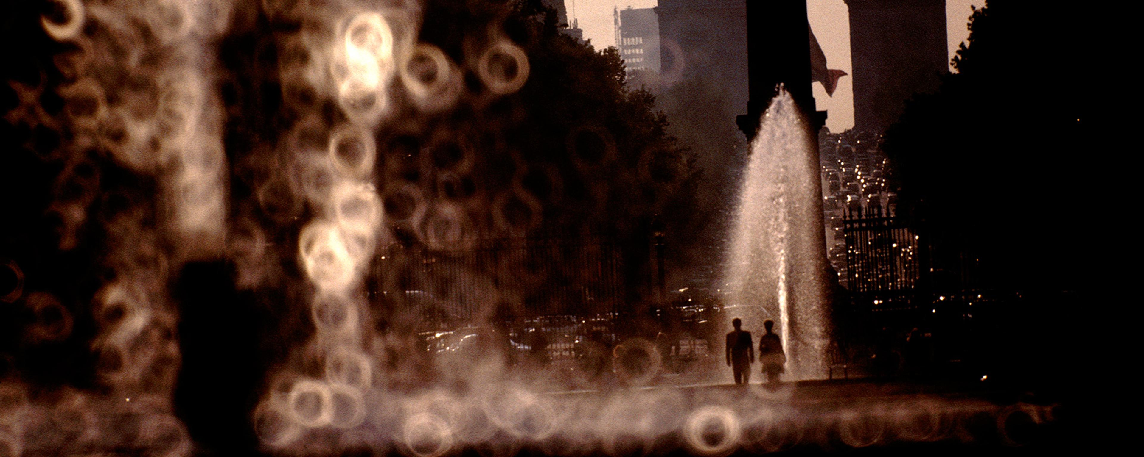 Photo of two silhouettes walking by fountains with bokeh effect in the foreground, Arc de Triomphe in the background.