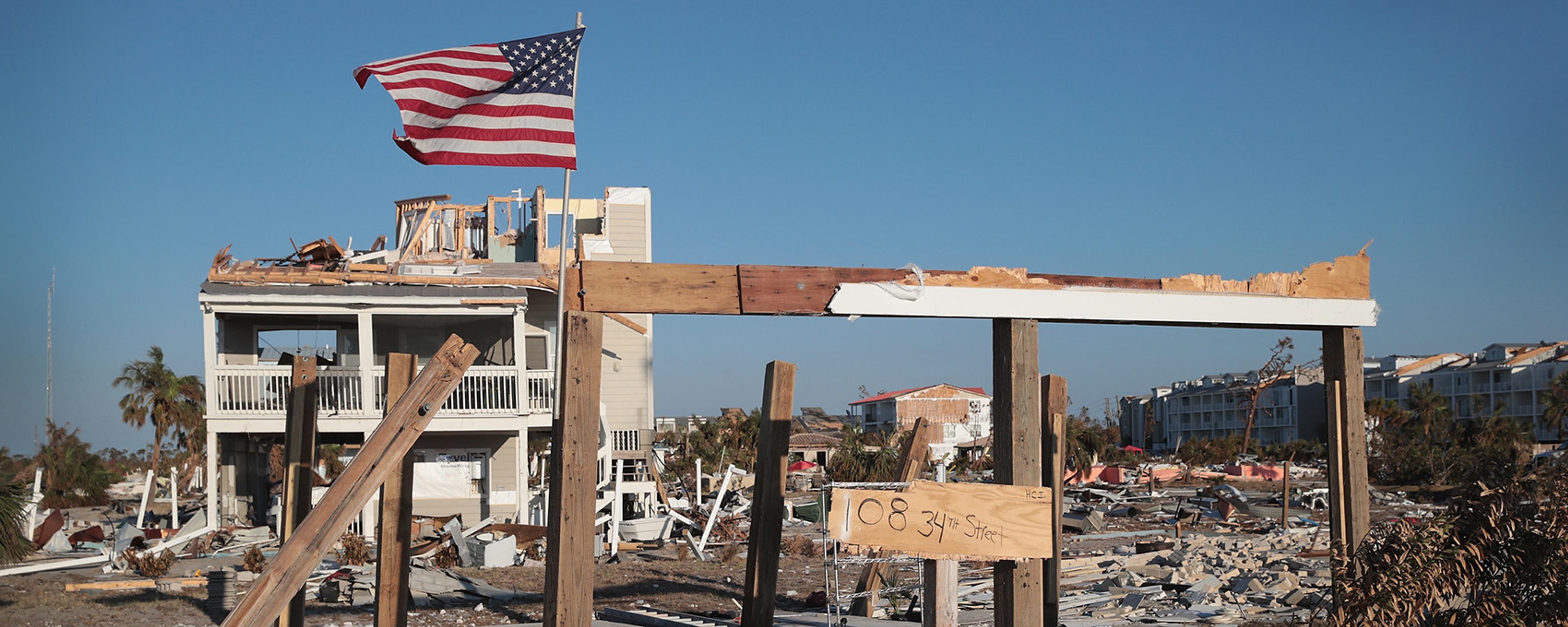 A destroyed house with an American flag amid debris and destruction under a clear blue sky.