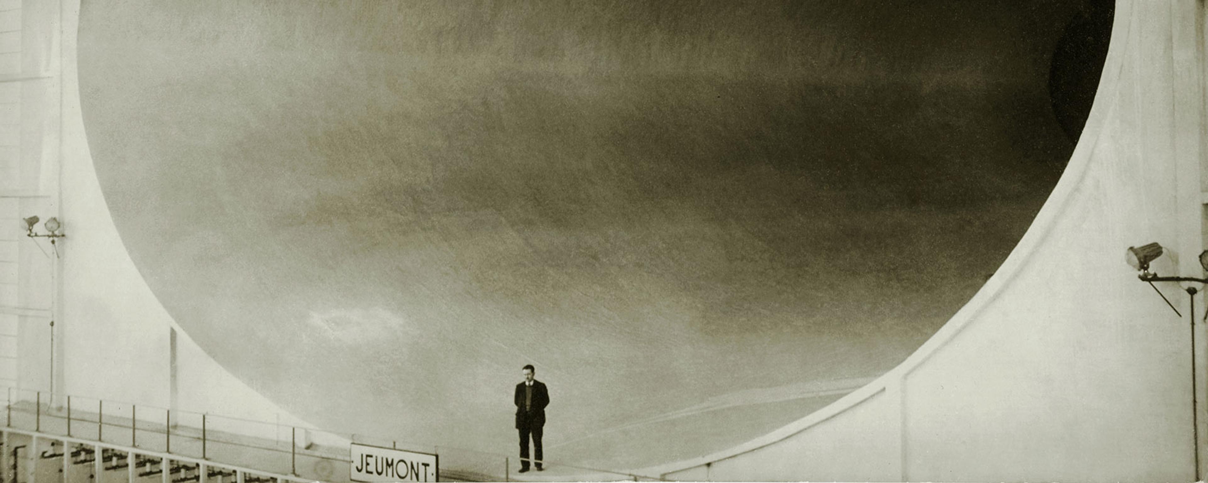 Black and white photo of man standing in front of a massive industrial tunnel with a Jeumont sign.