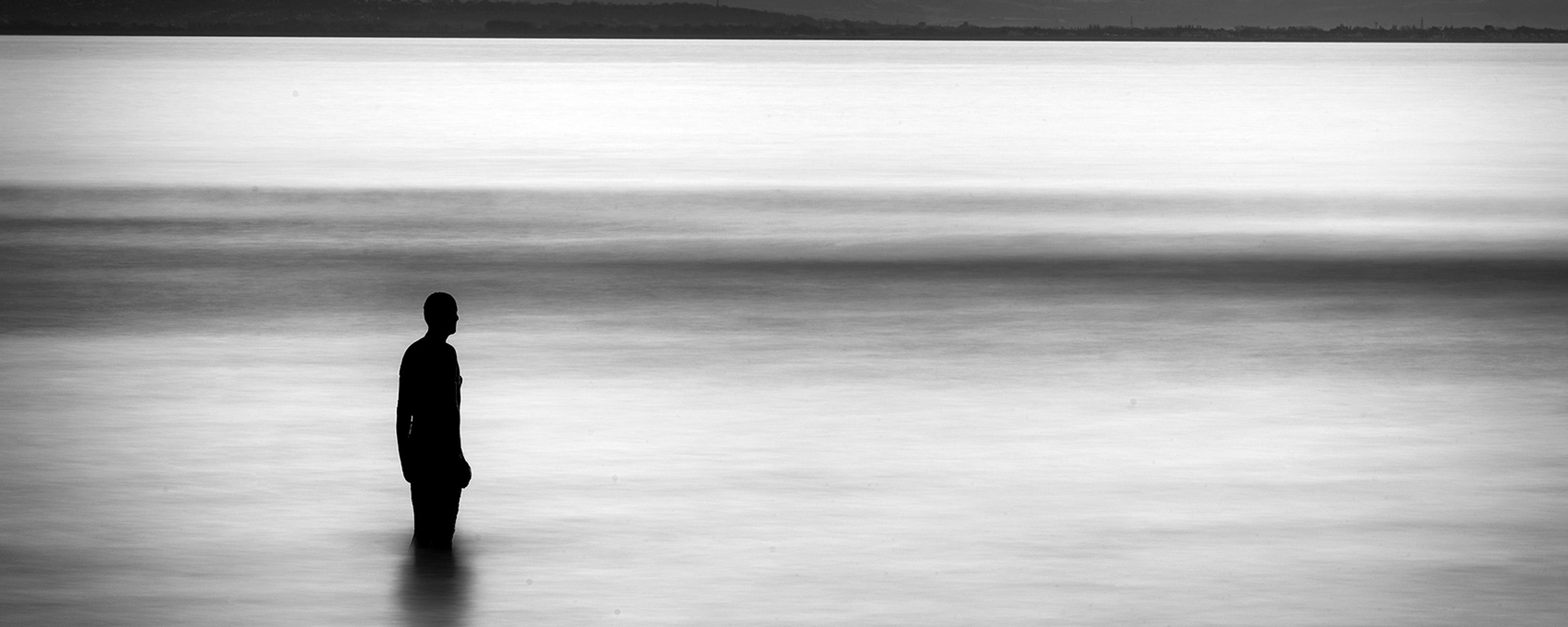 Black and white photo of a silhouette standing in calm water with distant hills and cloudy sky in the background.