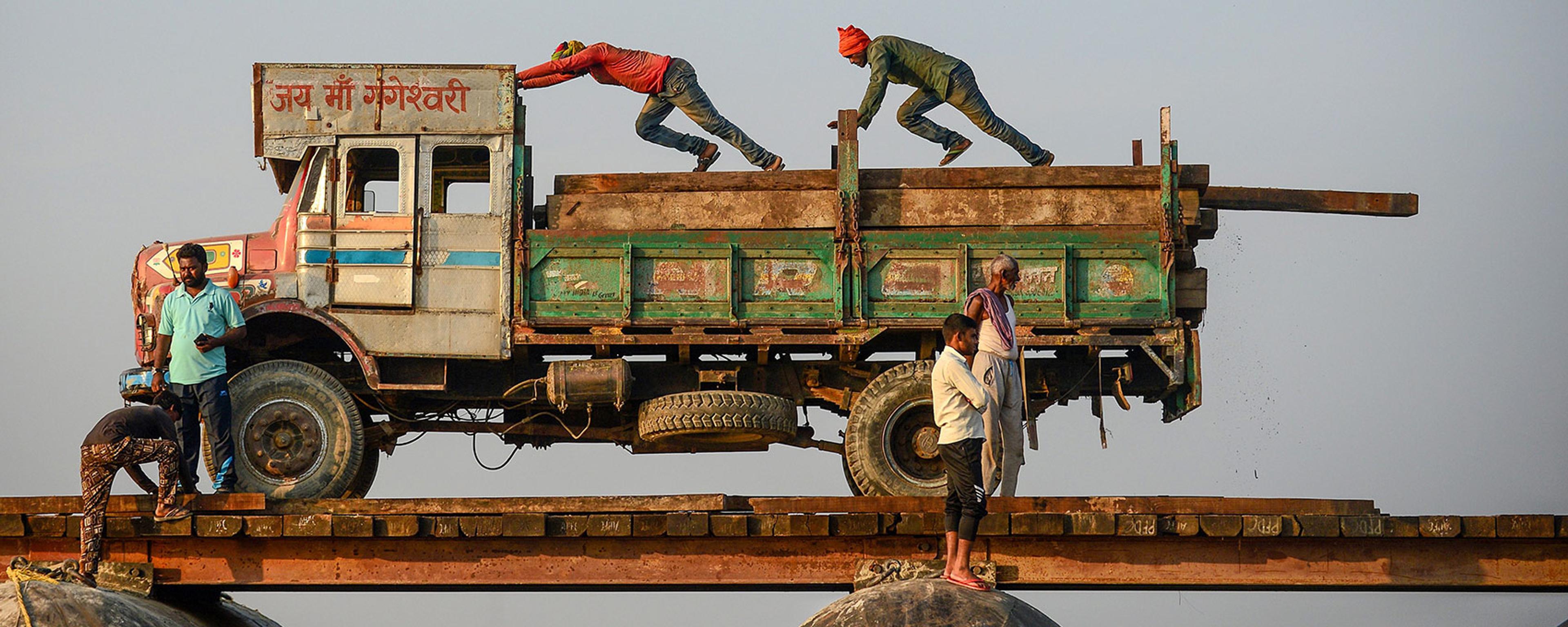 Photo of a truck on a narrow bridge with people balancing and pushing it under a clear sky.