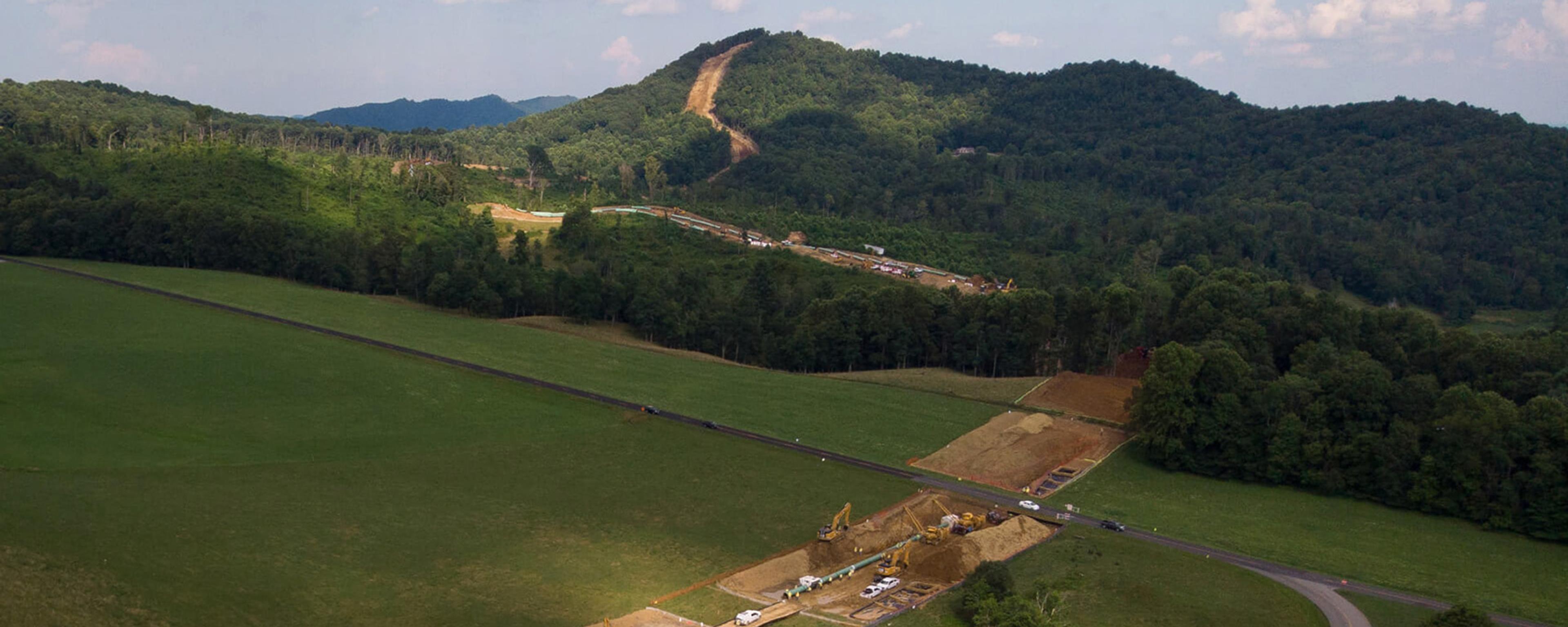 Aerial view of a large pipeline construction site with machinery and vehicles cutting through green fields and hills under a partly cloudy sky.