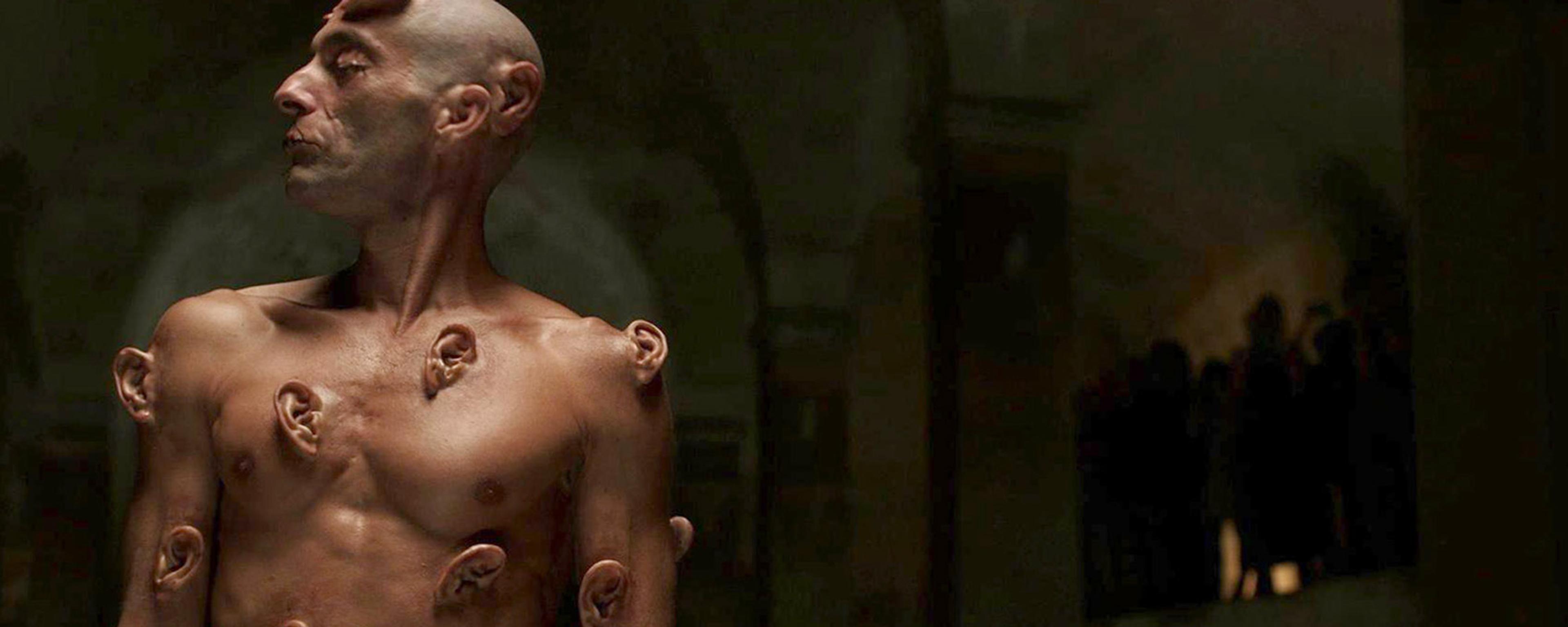 A shirtless man with multiple ears growing out of his body in strange places, in a dimly lit room.