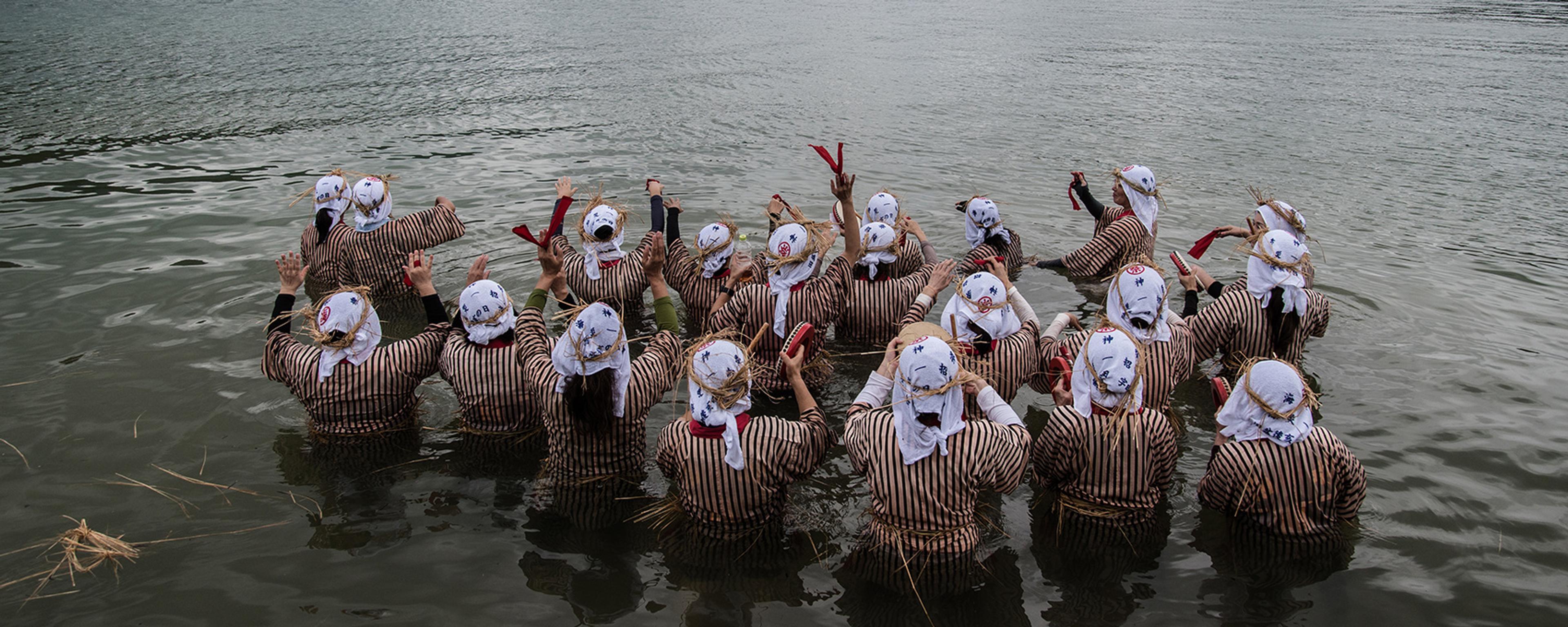 A group of people in striped clothing and headscarves standing in shallow water, raising hands towards a cloudy sky.