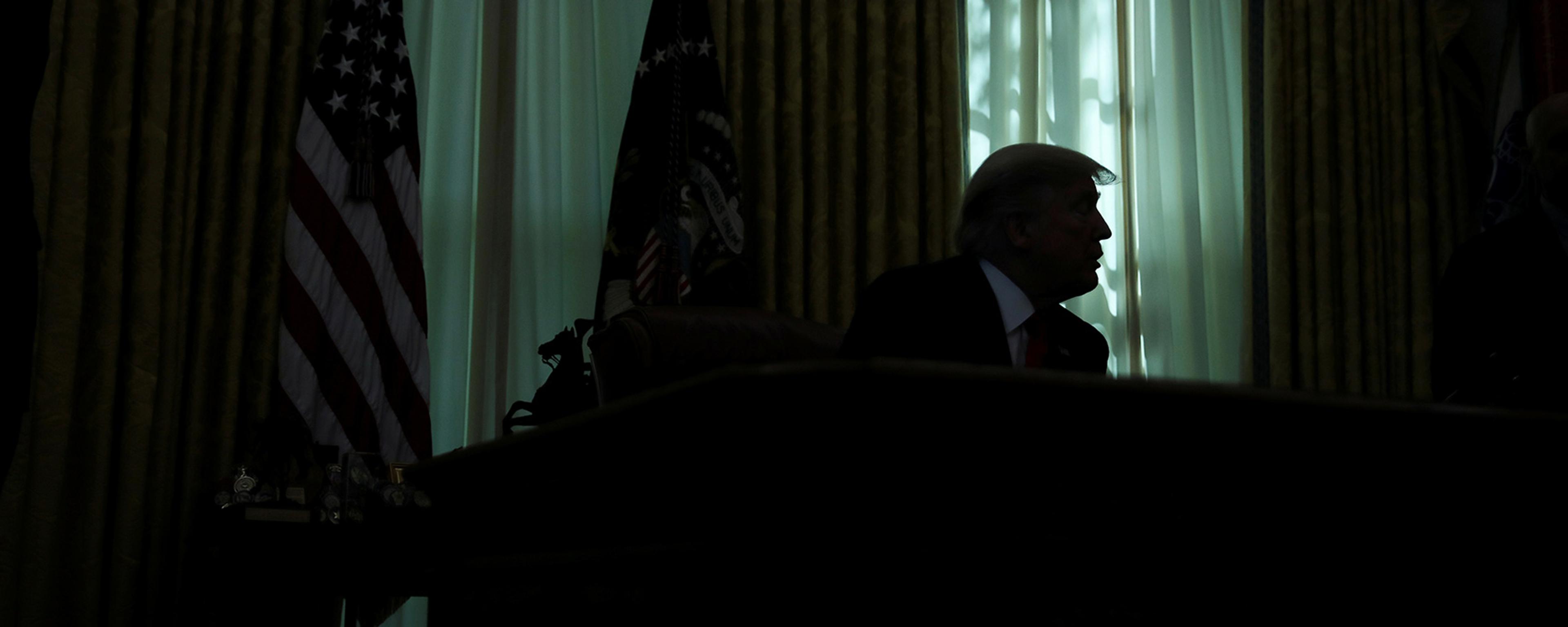 Silhouetted figure at a desk with US flag nearby, backlit by sunlight through curtains.