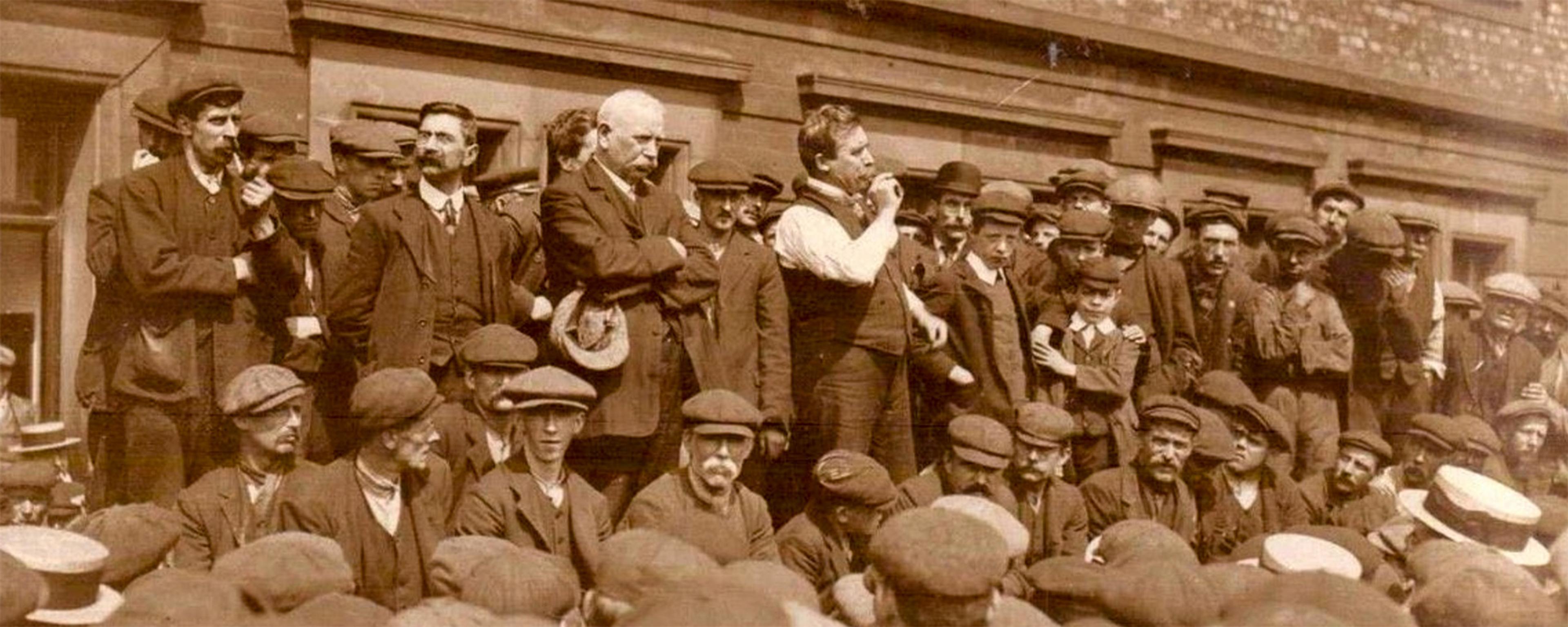 Sepia-toned photo of a man addressing a crowd of strikers outside a brick building wearing early 20th-century attire.