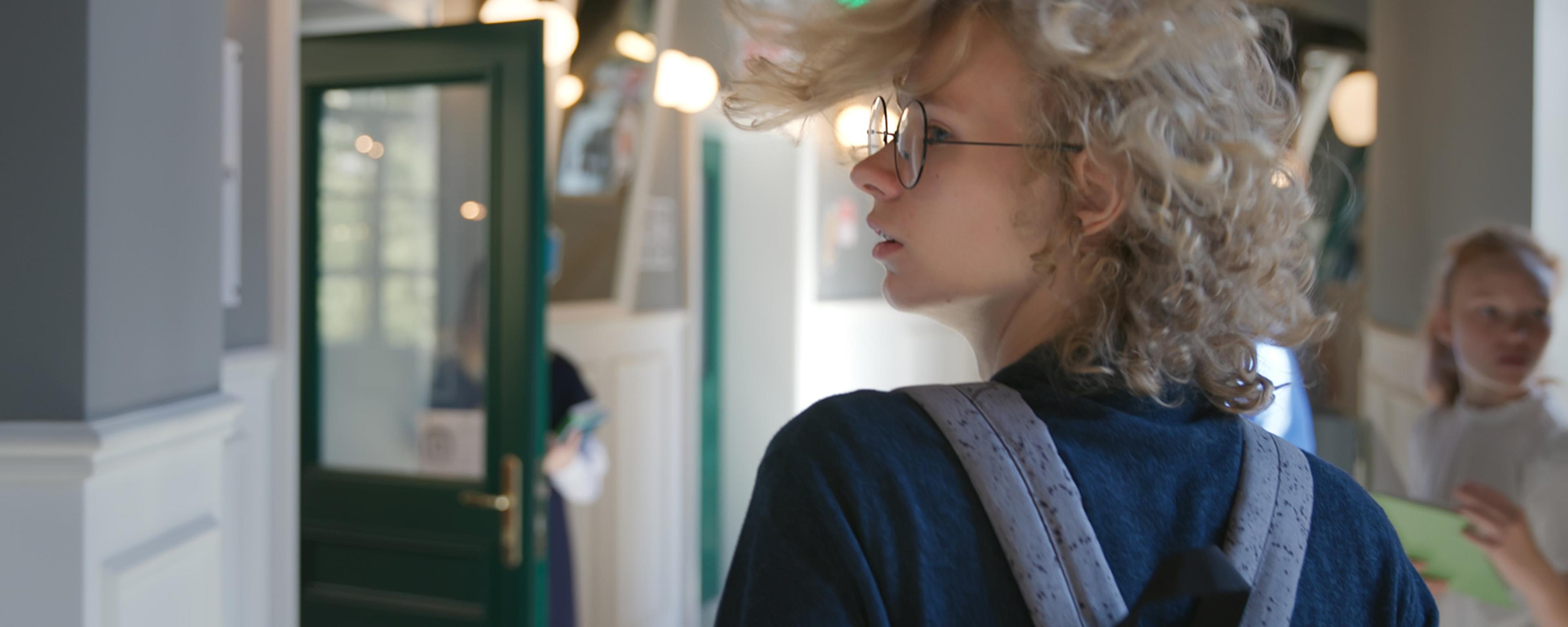 A young person with curly hair and glasses, wearing a backpack, indoors in what appears to be a school setting, with a green door and soft lighting, other children in background.